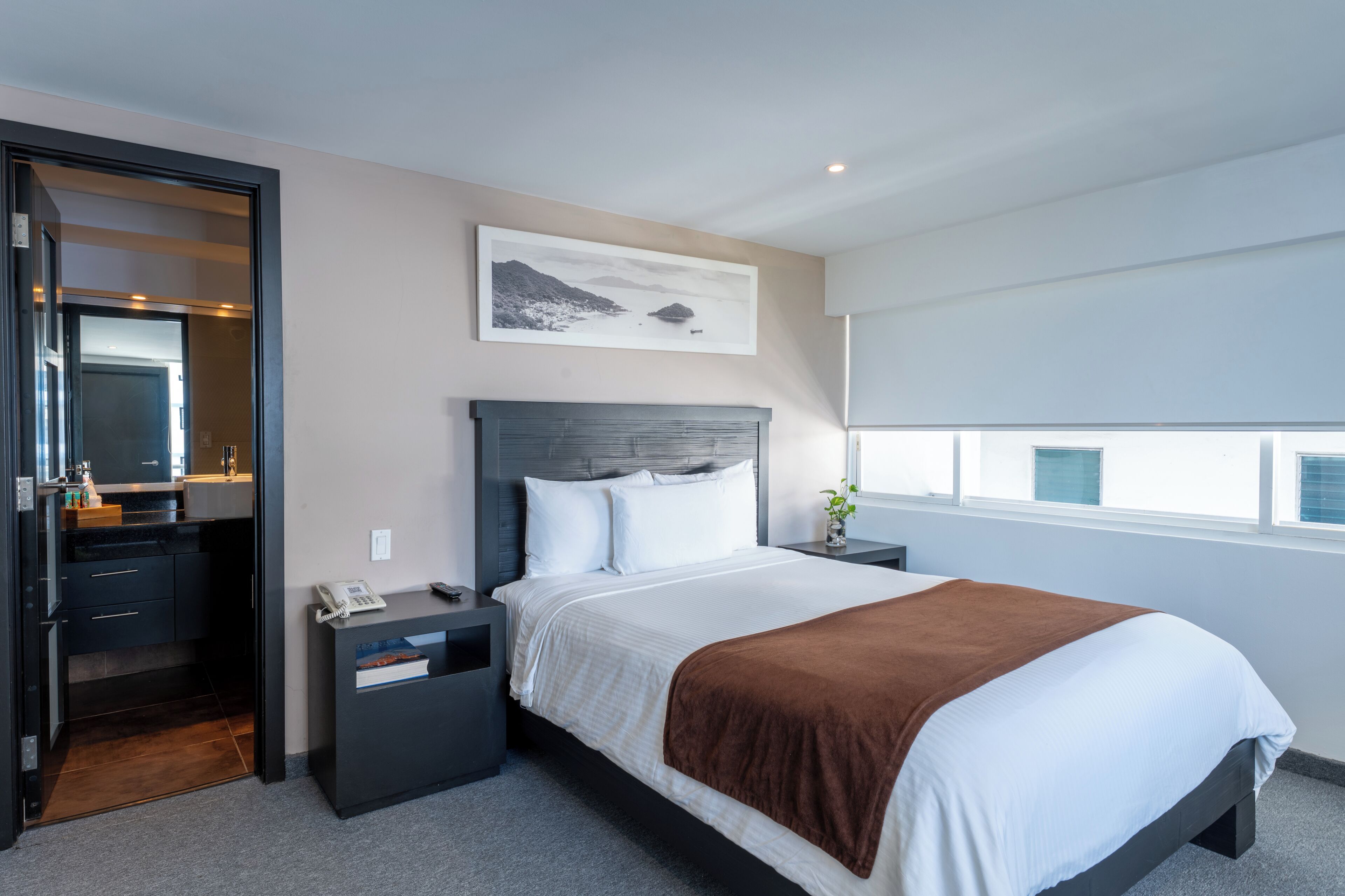 Habitación de hotel moderna con cama, mesitas de noche, escritorio con teléfono, espejo y ventana con persianas.