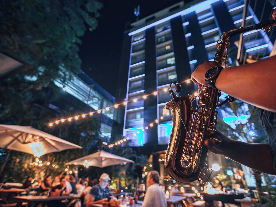 An outdoor dining area with tables and umbrellas, a person playing a saxophone, and a building with illuminated windows in the background.