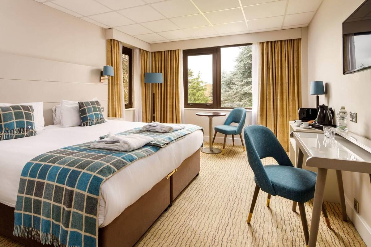 Habitación de hotel con cama, escritorio, silla, mesita y ventana con cortinas.