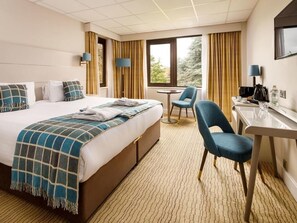 Quarto de hotel com cama, escrivaninha, cadeira, mesinha e janela.