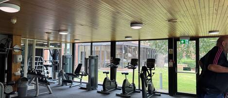 A gym with various exercise equipment, large windows, and a wooden ceiling.