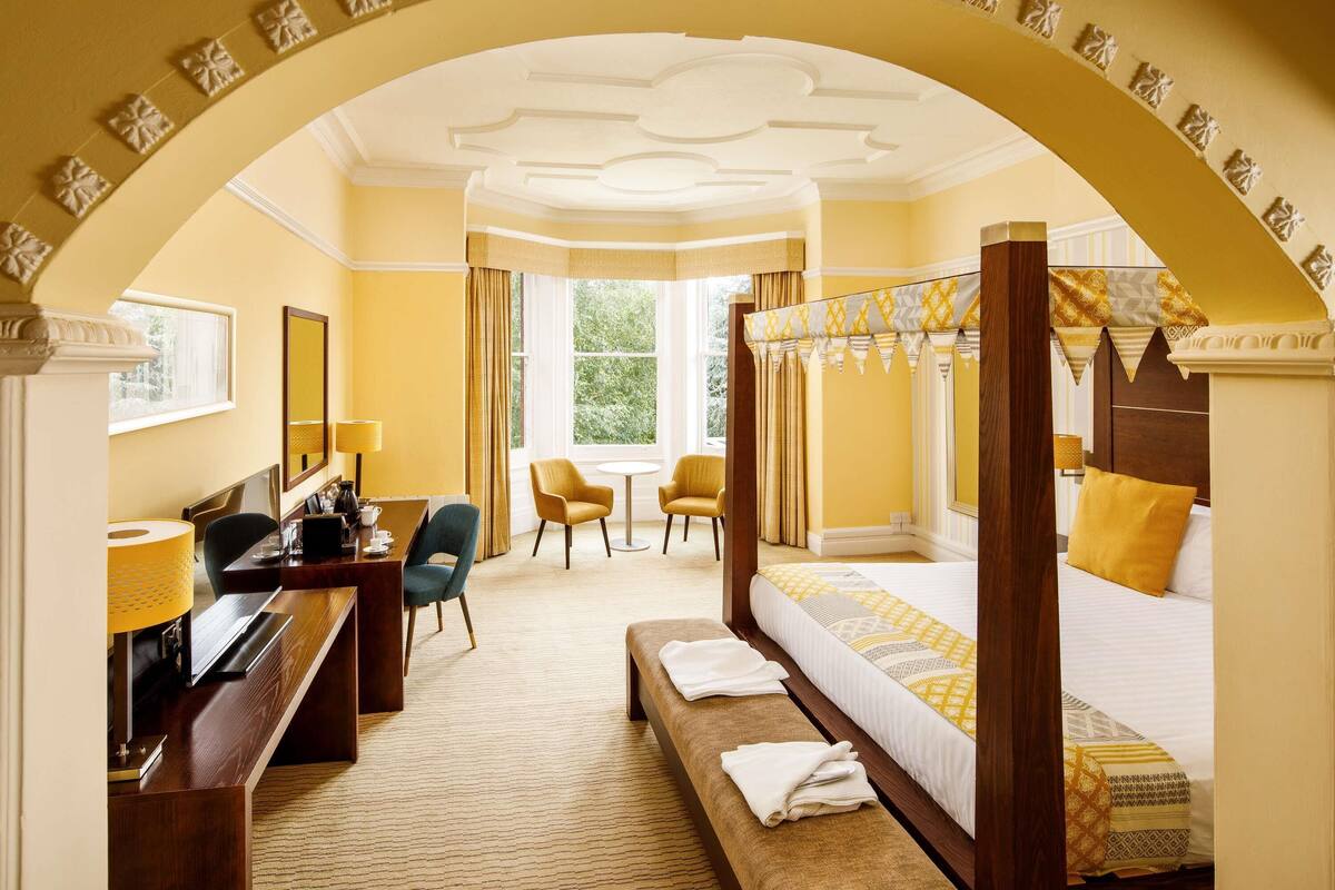 Une chambre d’hôtel avec un lit à baldaquin, un bureau avec une chaise, une petite table et une fenêtre avec des rideaux.