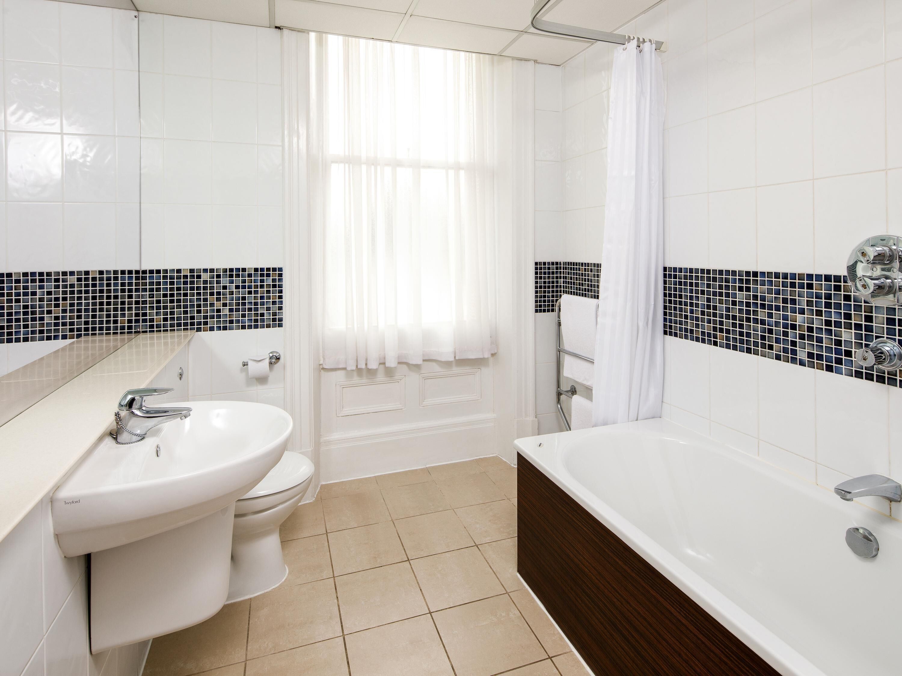 A bathroom with a white sink, toilet, and bathtub, tiled walls, and a window with a white curtain.