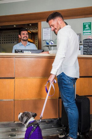 A hotel reception area with a counter, a man in a white shirt and jeans, and a dog on a leash.