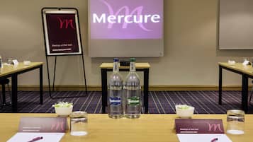 A conference room with a presentation screen displaying the word 'Mercure,' two tables with water bottles, nameplates, pens, and a small bowl of salad.