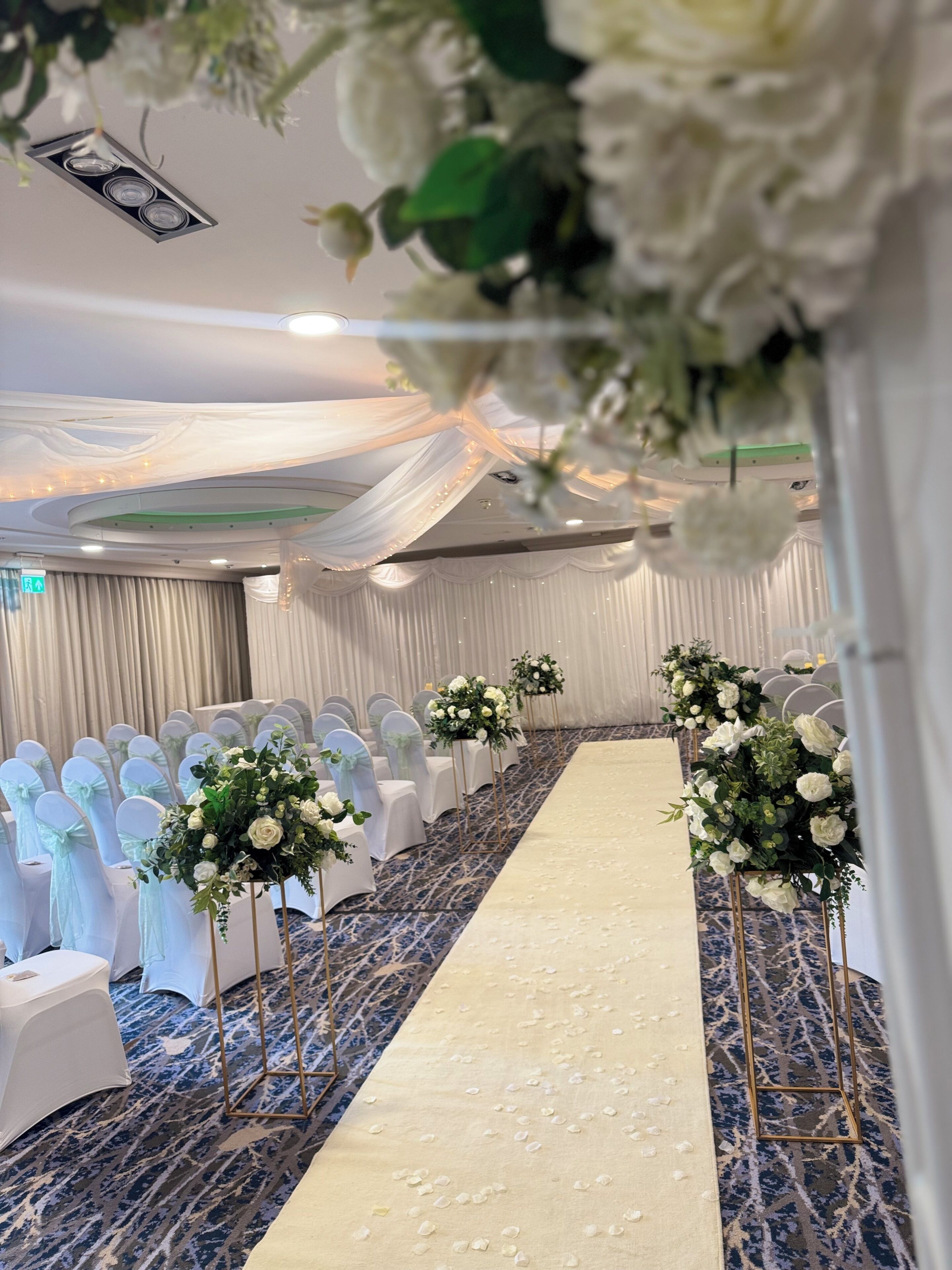 Un salón de banquetes con filas de sillas blancas, un pasillo central y centros de mesa florales.