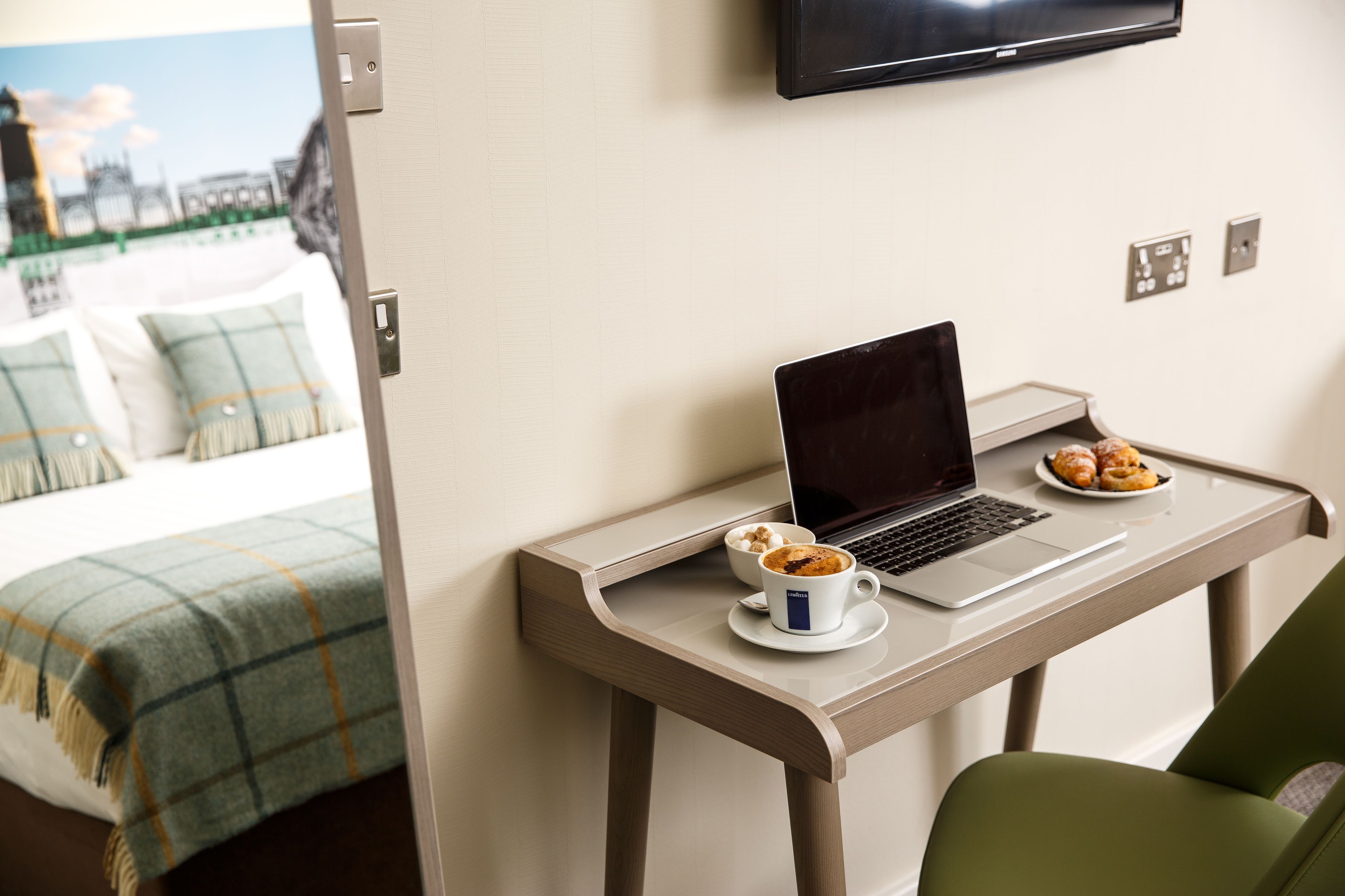 Een hotelkamer met een bed, een bureau met een laptop, een kop koffie en een bord gebak.