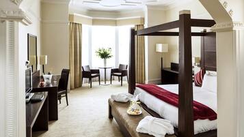 Habitación de hotel con cama con dosel, un escritorio con computadora, una silla, una mesita con un jarrón de flores y vistas al exterior a través de amplias ventanas.