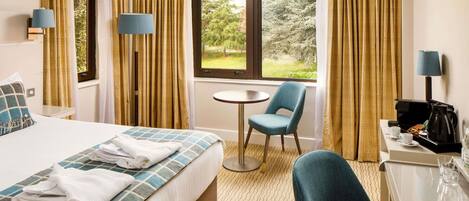 Habitación de hotel con cama, escritorio, silla, cafetera y ventana con vista a árboles.
