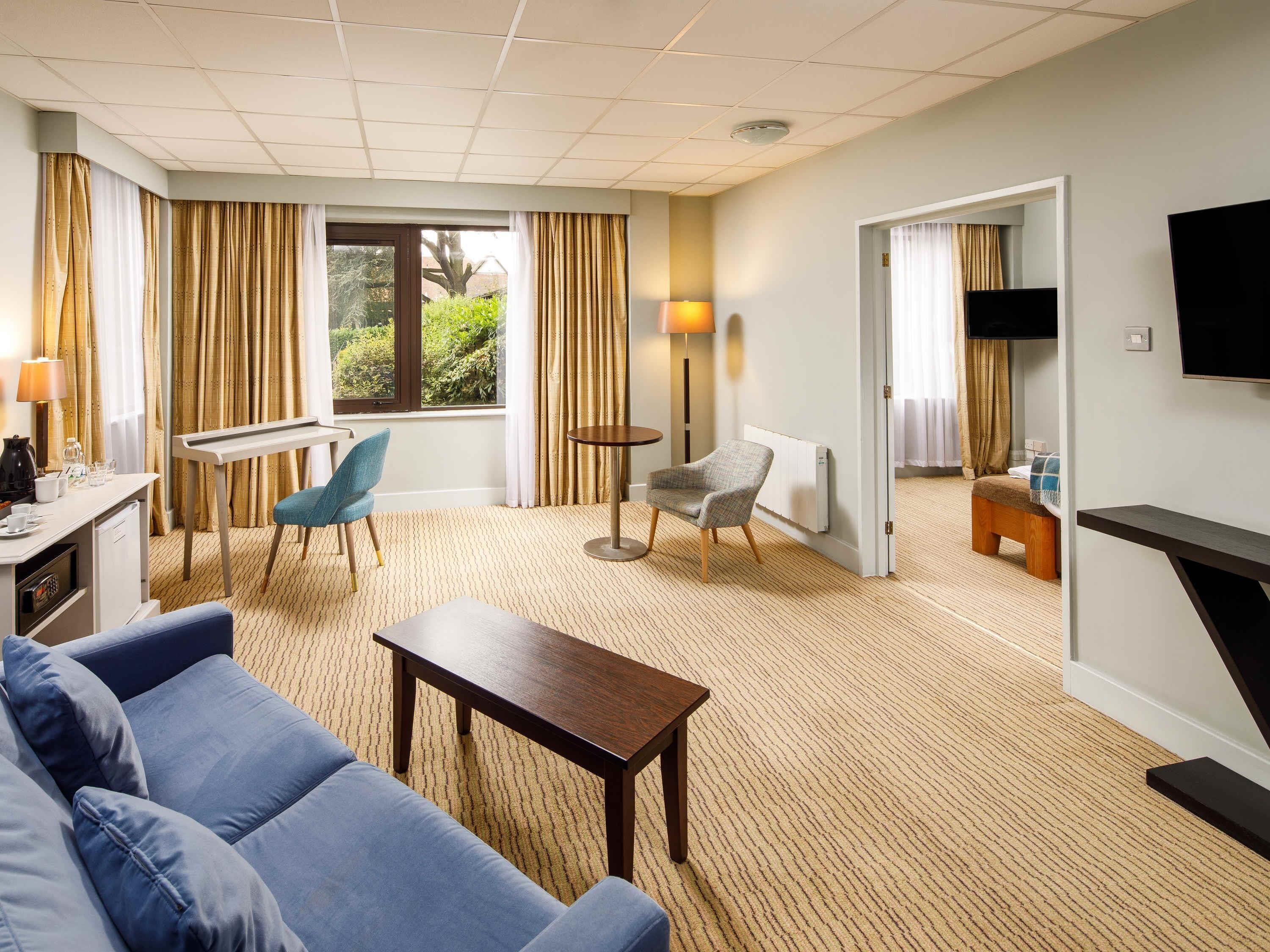 A hotel room with a sofa, a small table, a chair, a TV, a desk with a lamp, and a view of greenery outside.