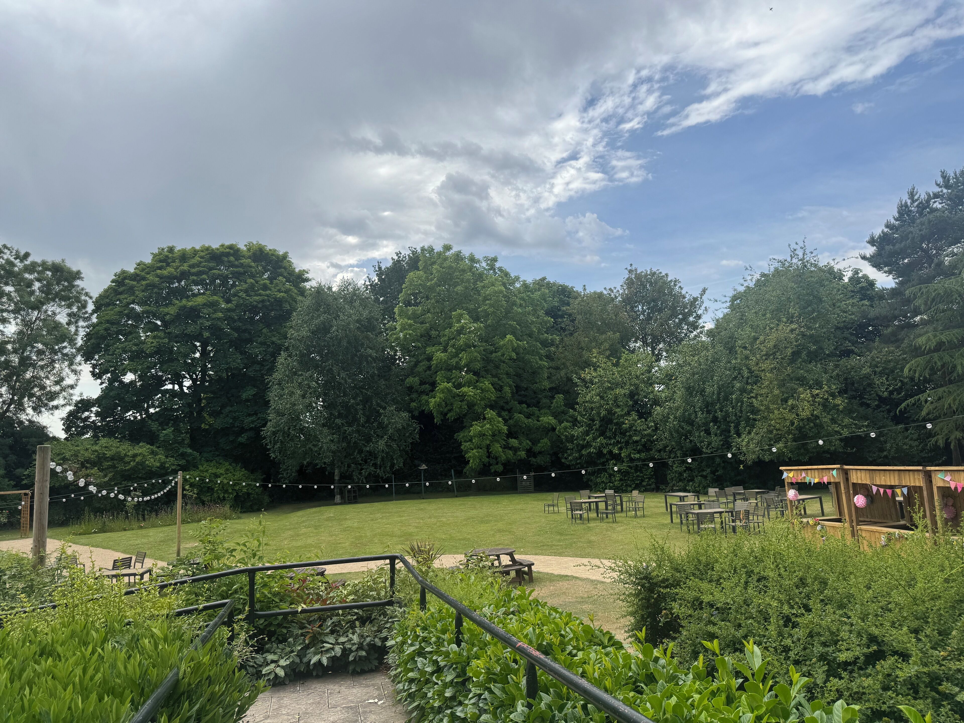 A grassy outdoor area with picnic tables and a wooden structure, surrounded by dense trees.