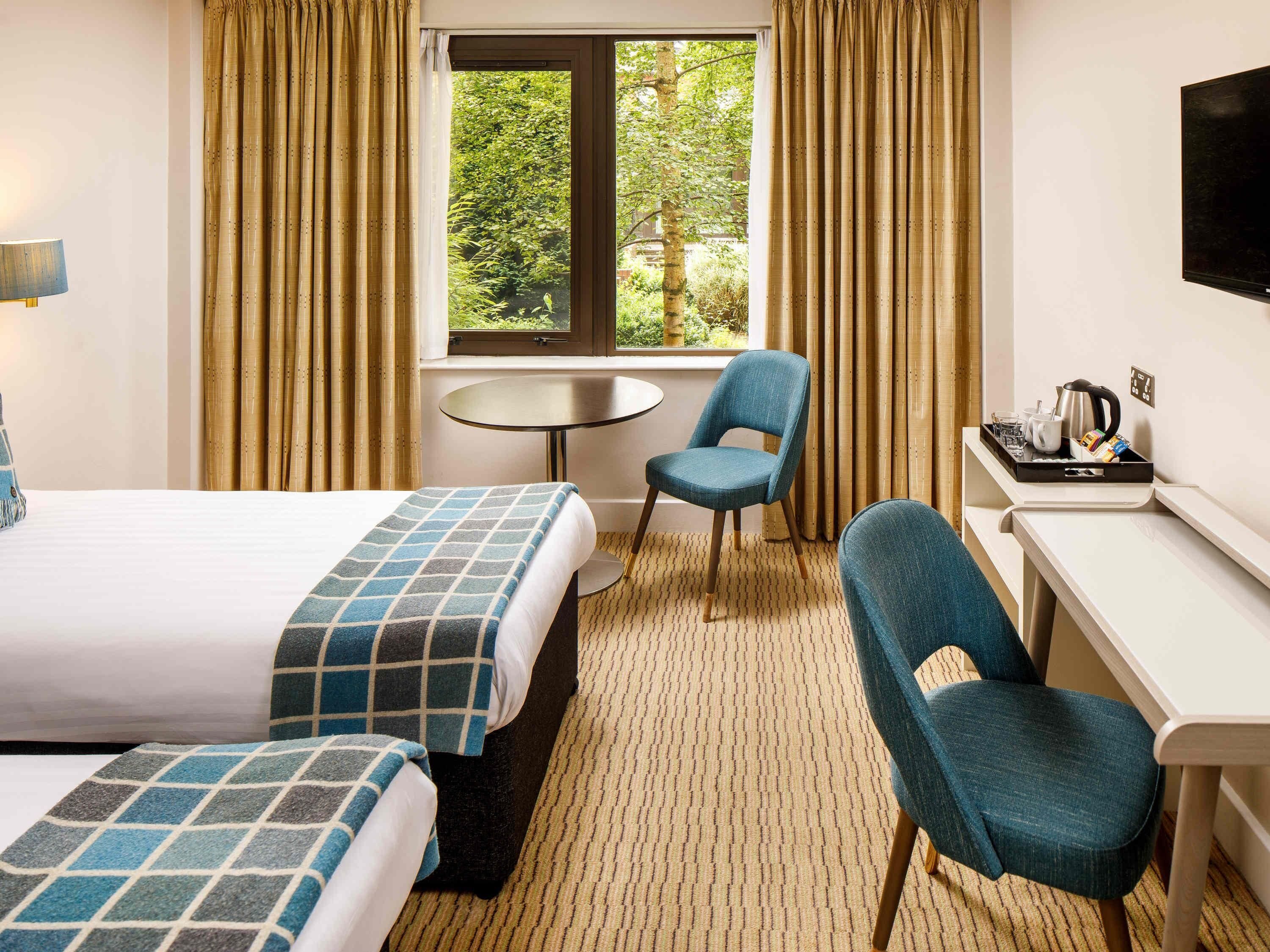A hotel room with two beds, a desk, a chair, a small table, and a window with curtains.
