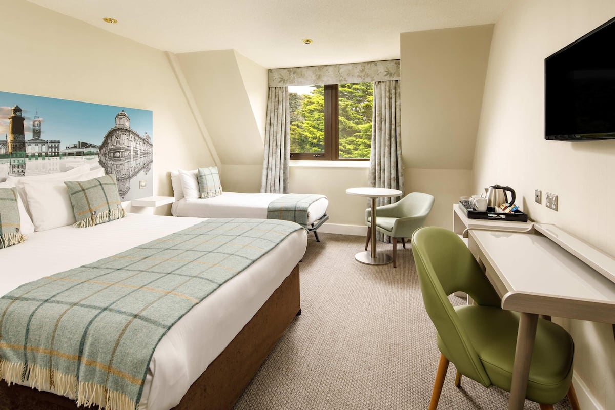 Une chambre d’hôtel avec un grand lit, un lit plus petit, un bureau avec une chaise, une table ronde et un téléviseur fixé au mur.