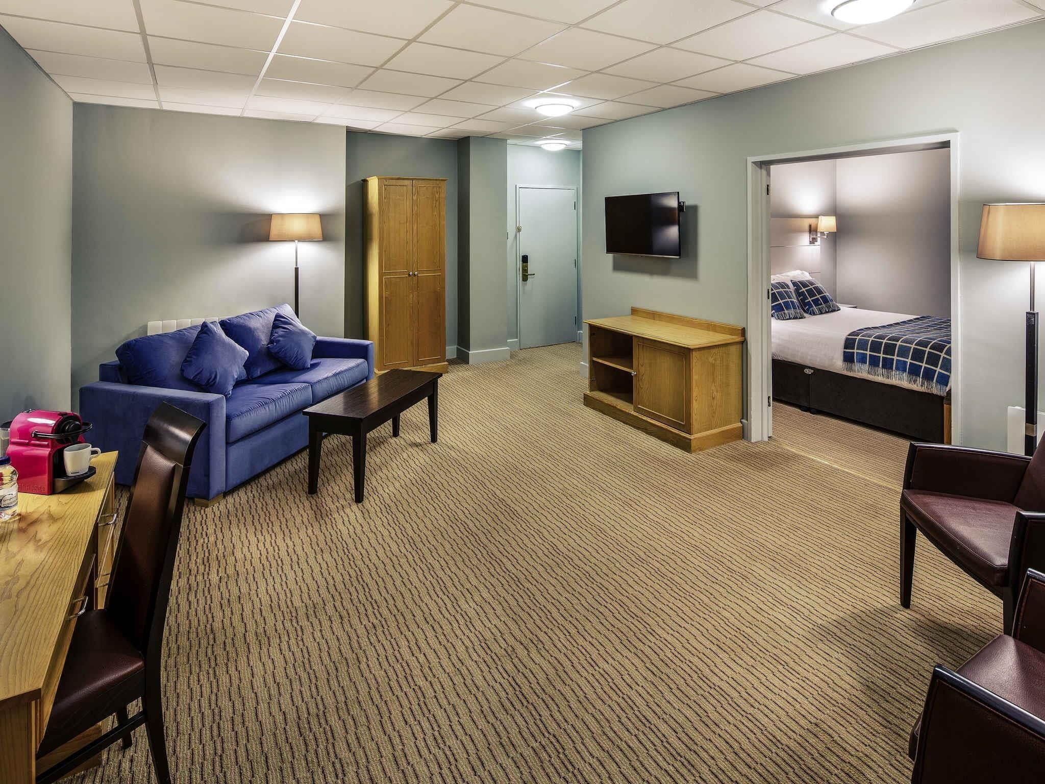 A hotel room with a blue sofa, a wooden cabinet, a bed with a blue and white checkered blanket, a television mounted on the wall, and a small table with a coffee maker.