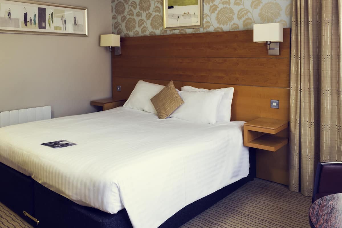 Une chambre d’hôtel avec un grand lit, une tête de lit en bois, deux tables de chevet avec des lampes, un tableau encadré au mur et un sol recouvert de moquette.