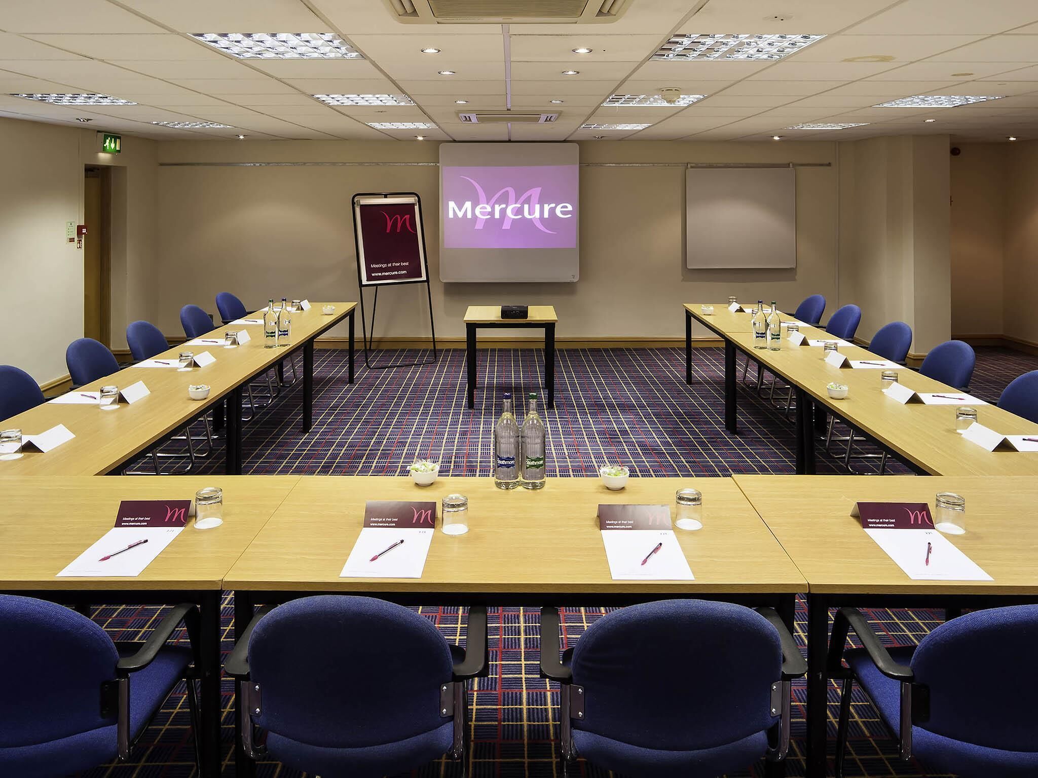 A conference room with a long table, blue chairs, nameplates, and a projector screen displaying 'Merture'.