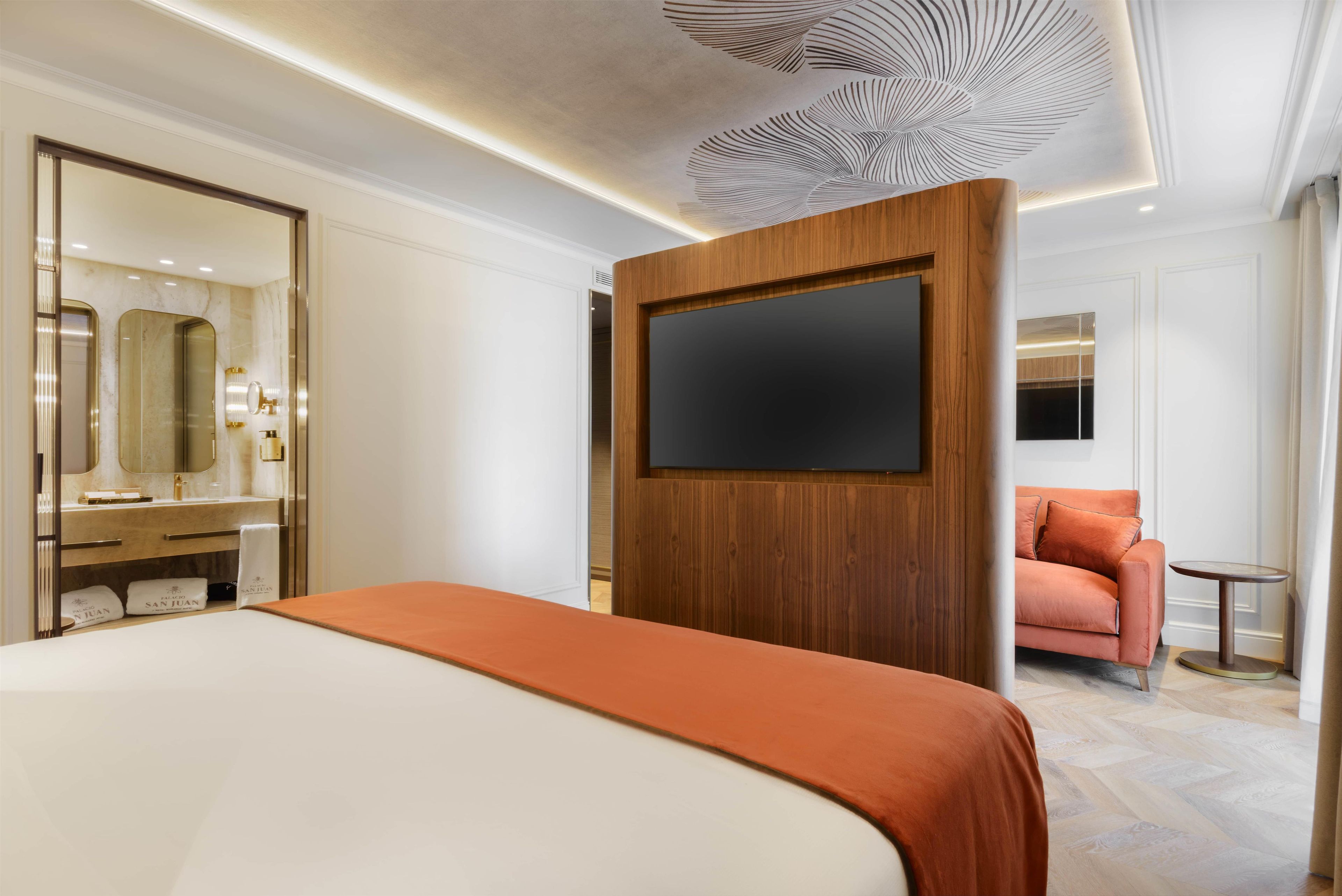 A hotel room with a bed, a TV mounted on a wooden panel, a bathroom with a mirror and vanity, and a seating area with a sofa and table.