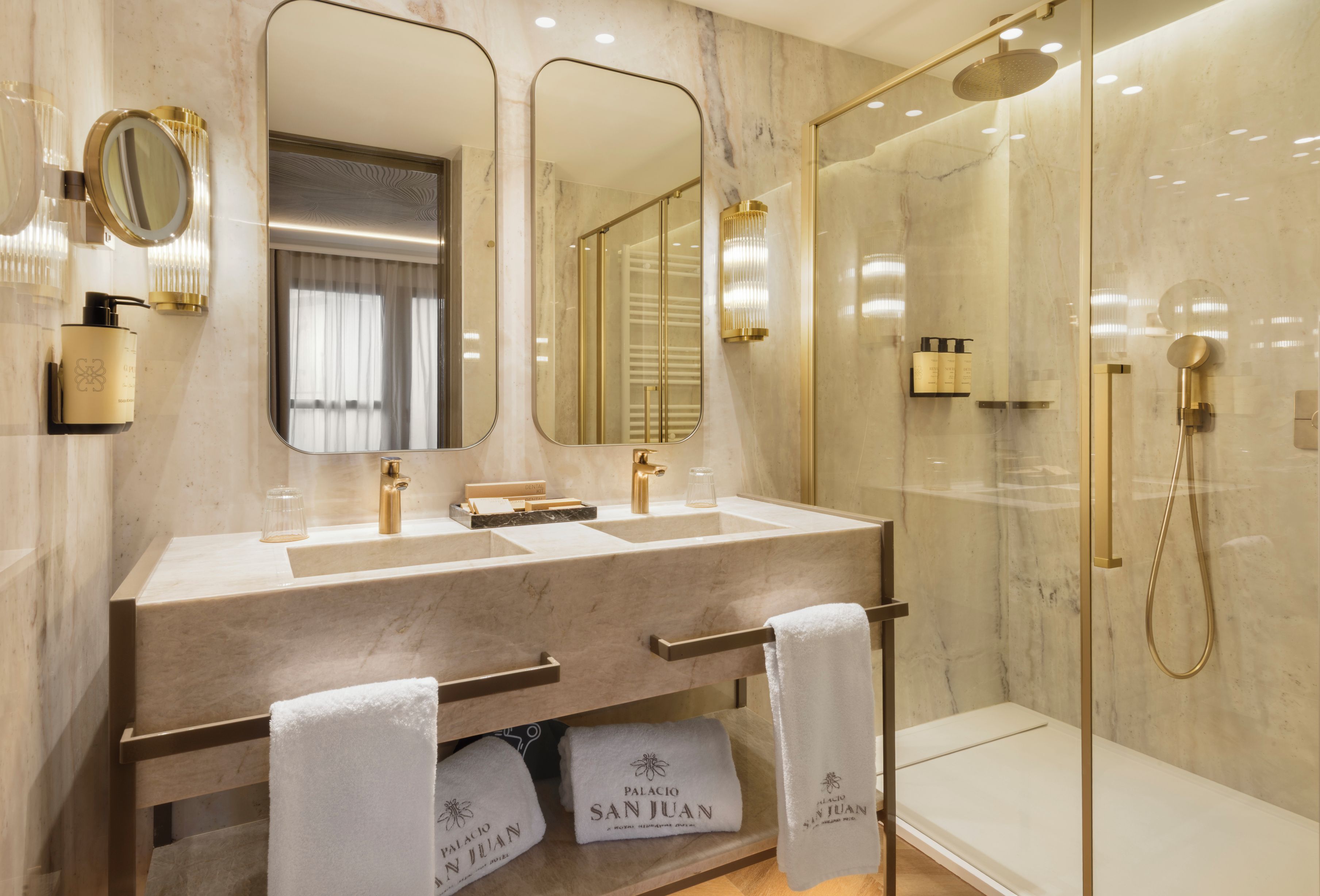 Un baño moderno con tocador de mármol, doble lavamanos, un espejo grande y zona de ducha con mampara de vidrio.