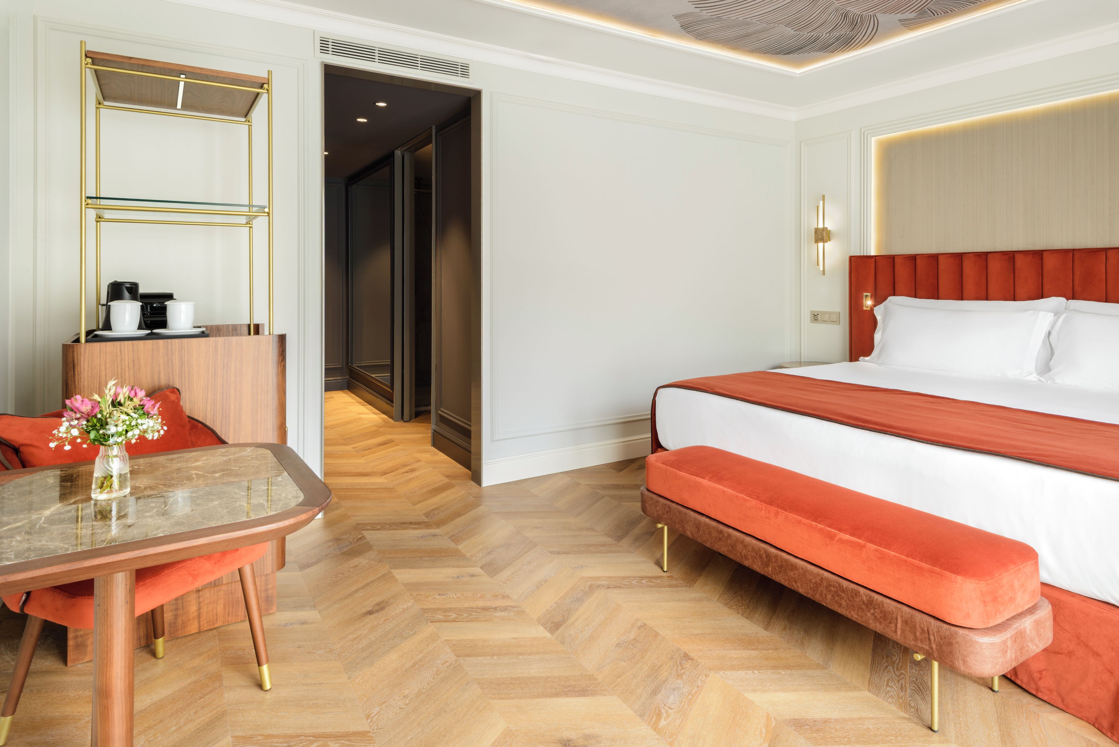 Habitación de hotel con cama, escritorio con cafetera, florero con flores y suelo de madera tipo espiga.