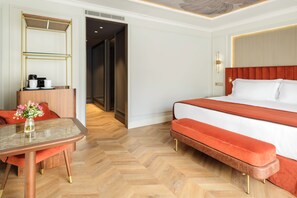 Habitación de hotel con cama, escritorio con cafetera, florero con flores y suelo de madera tipo espiga.