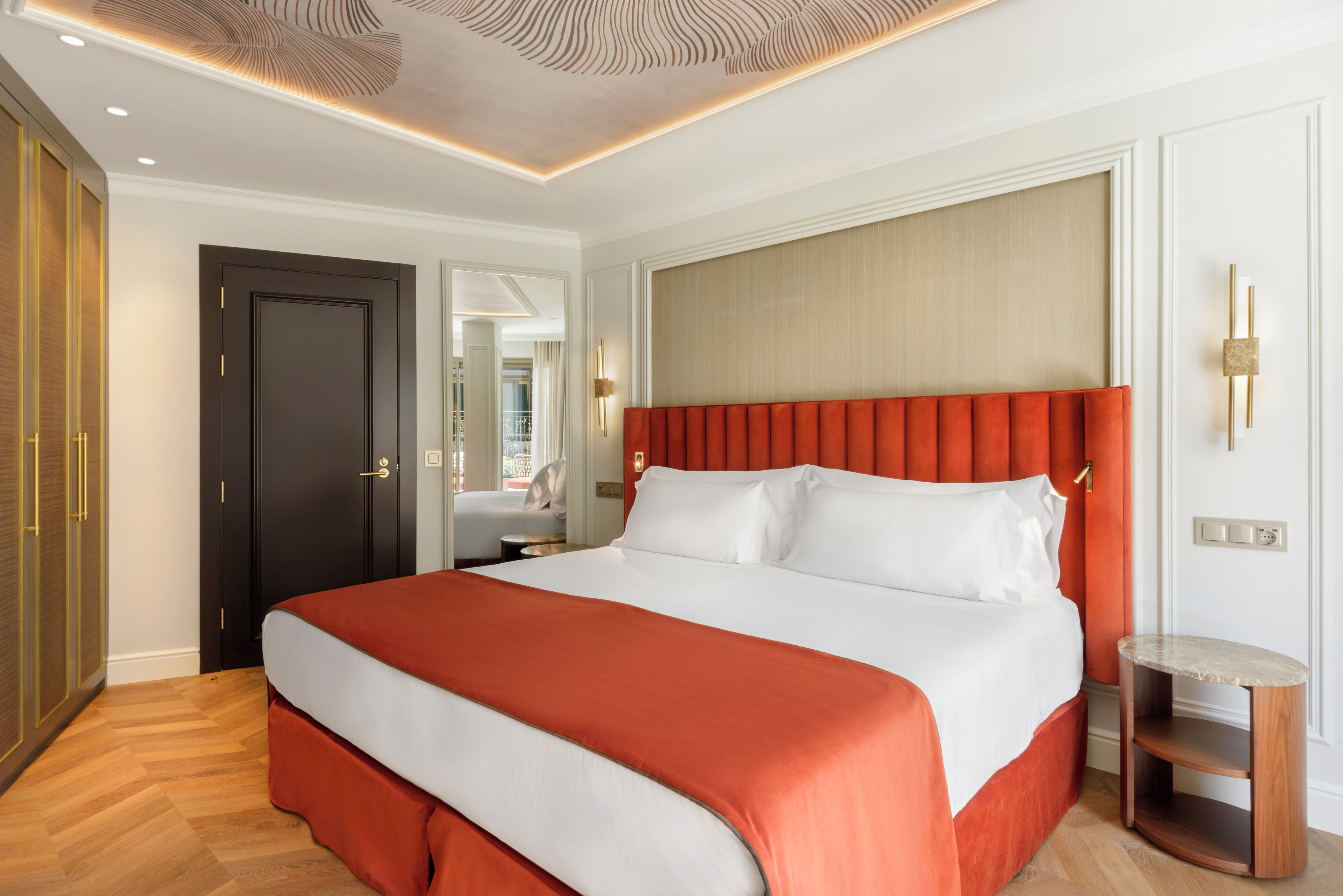 Een hotelkamer met een groot bed, een houten vloer, een donkere deur en een wandlamp.