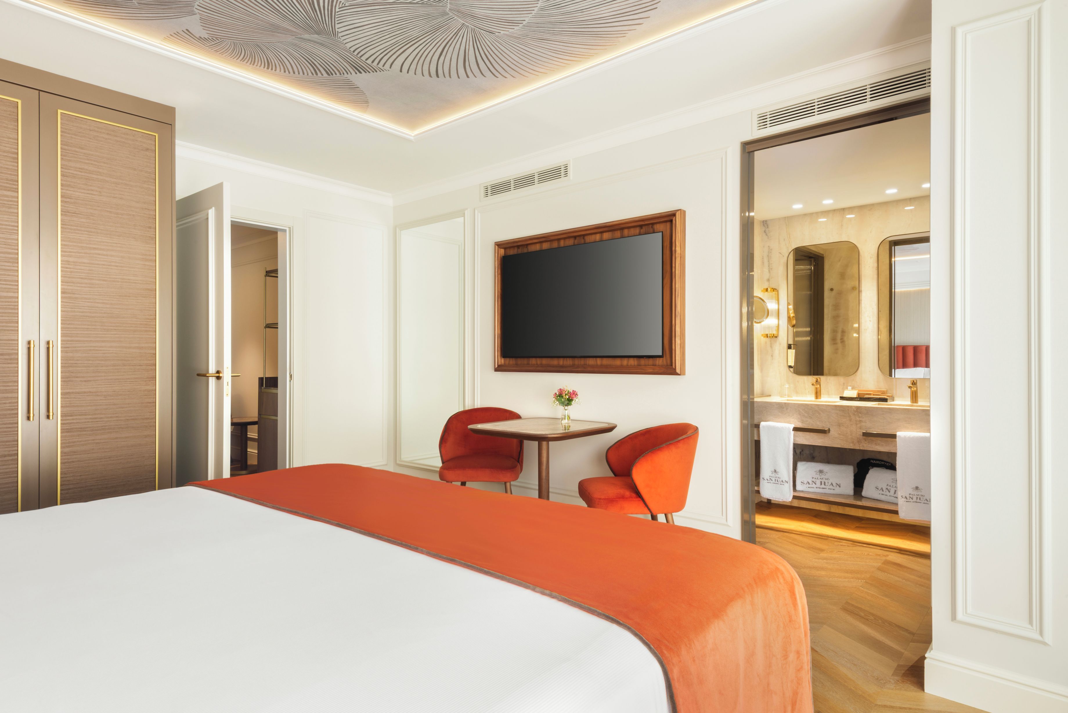 Habitación de hotel con una cama grande, un escritorio, dos sillas, un espejo y un baño visible a través de una puerta abierta.