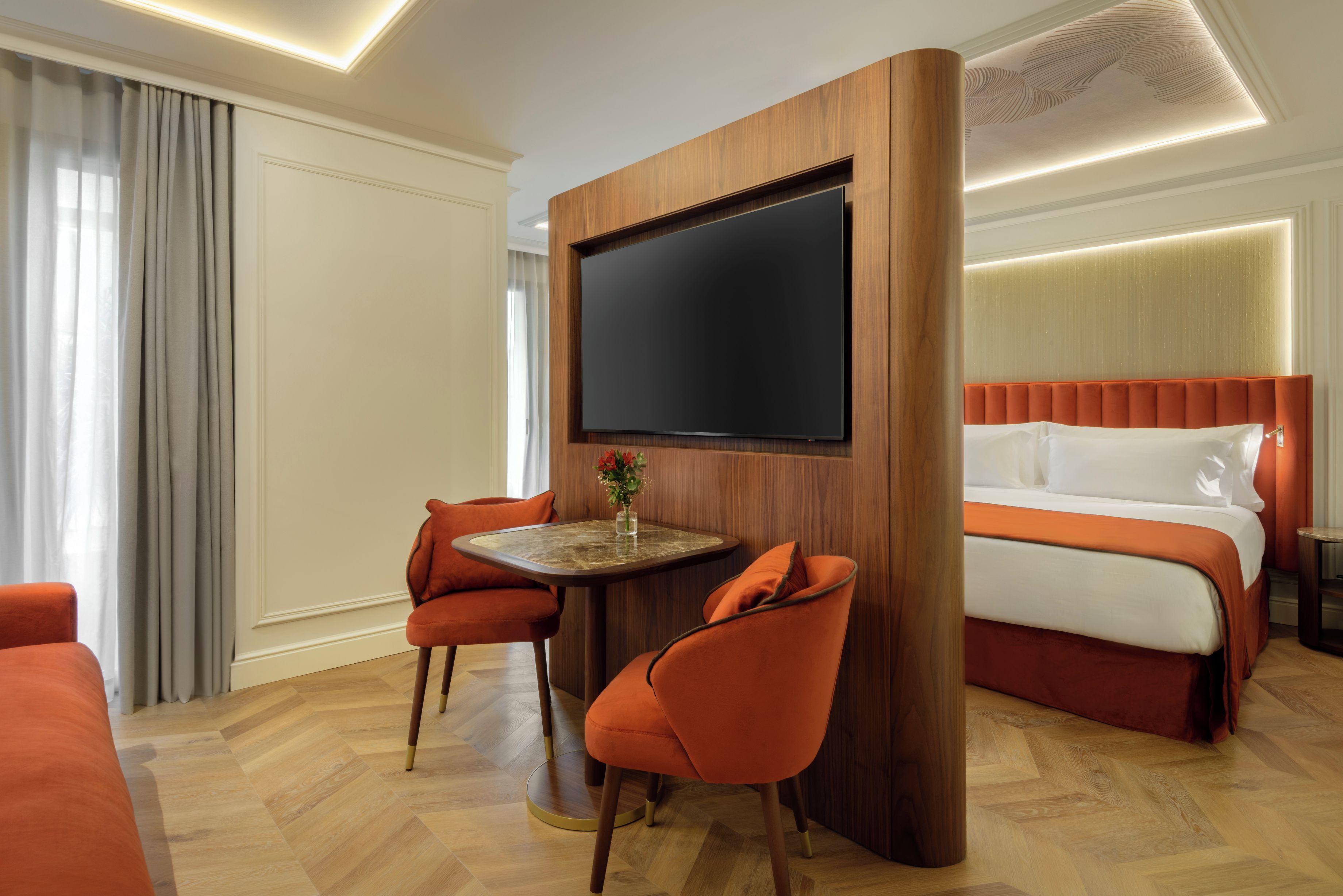 Een hotelkamer met een bed, een televisie, twee stoelen, een klein tafeltje en een raam met gordijnen.