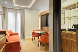 A hotel room with a sofa, a dining table, chairs, a TV, and a coffee machine.