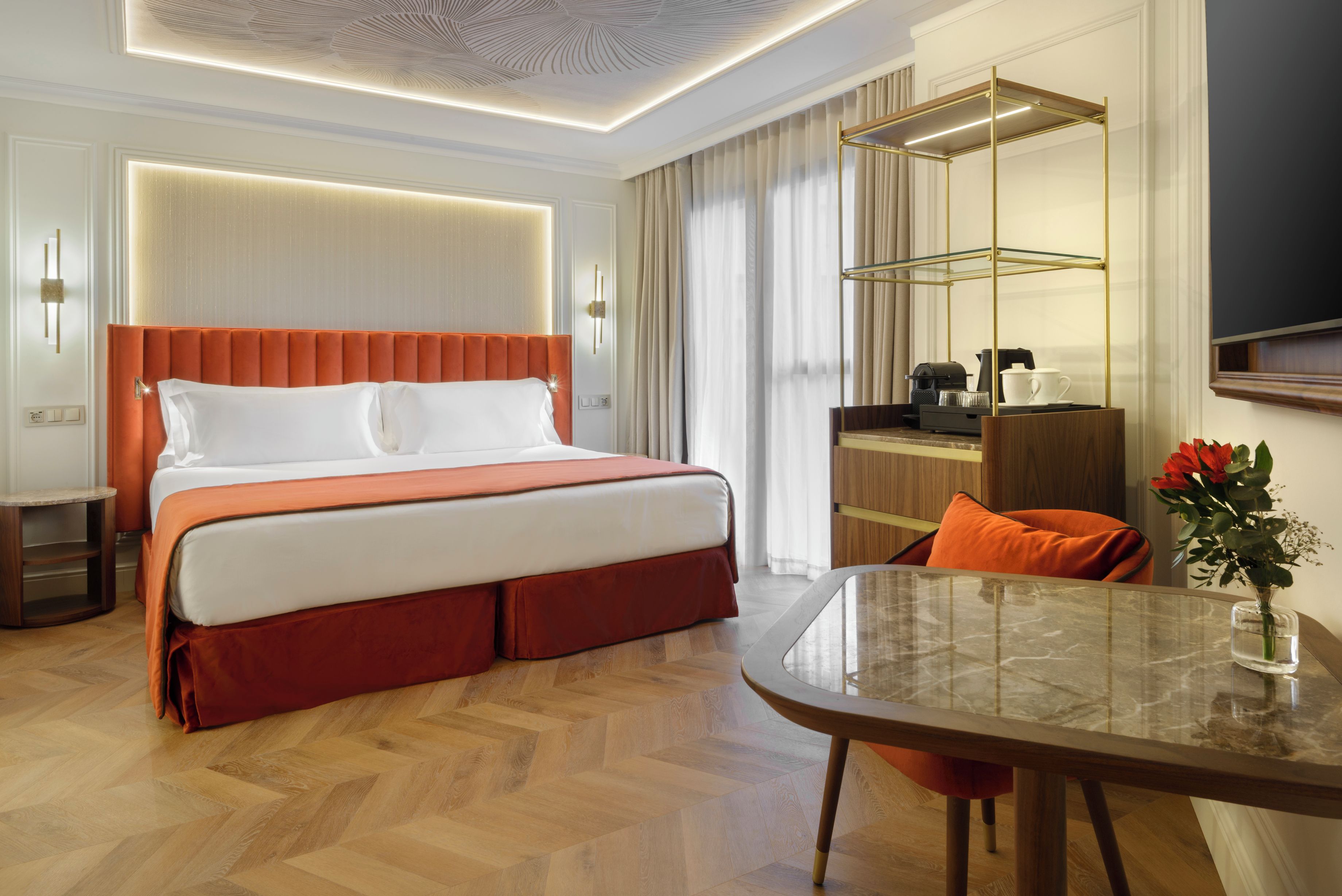 Habitación de hotel con una cama grande, un escritorio con silla, una mesa de centro de vidrio y un florero con flores.