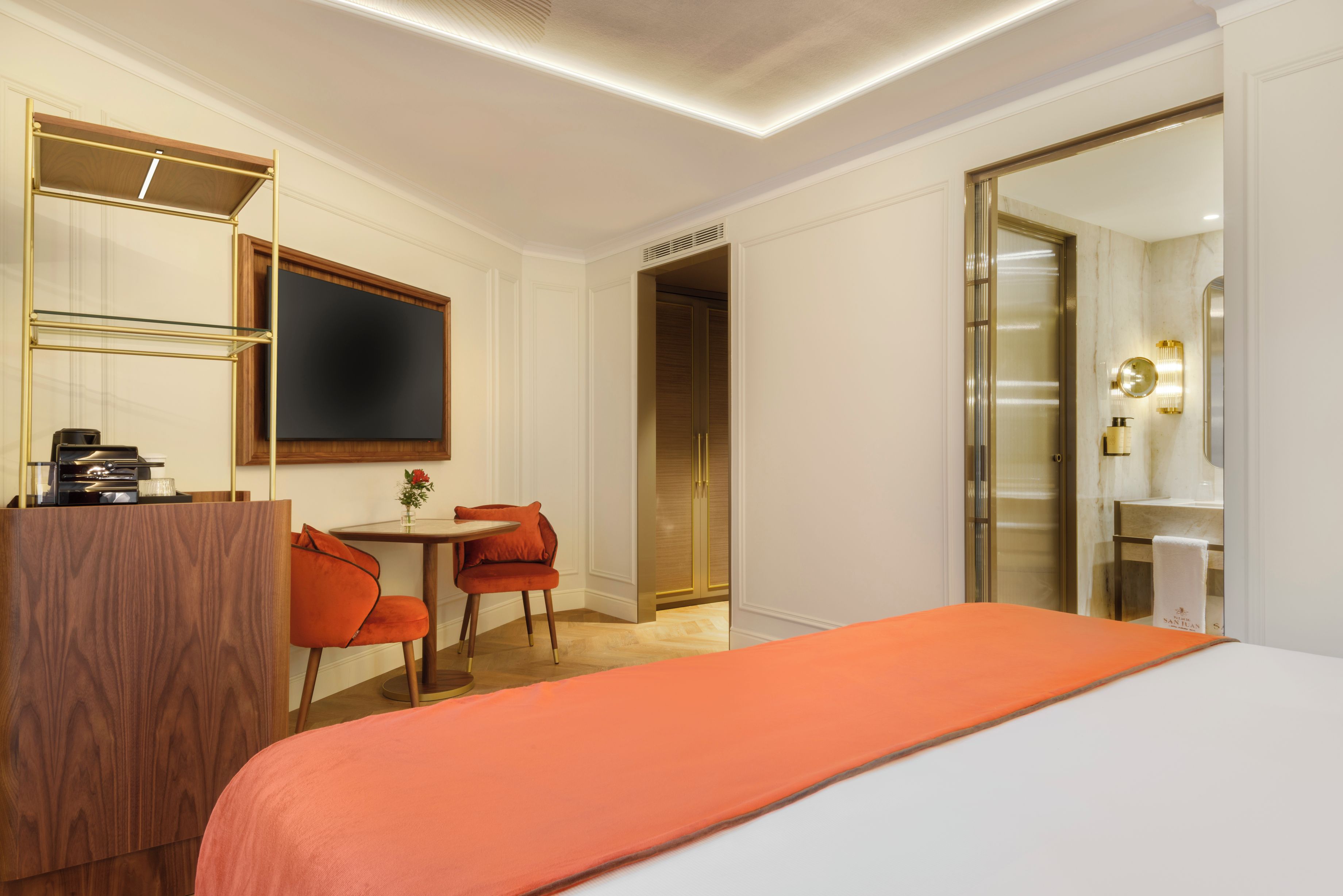 Habitación de hotel con cama, escritorio con silla, televisión y baño con espejo y lavamanos.