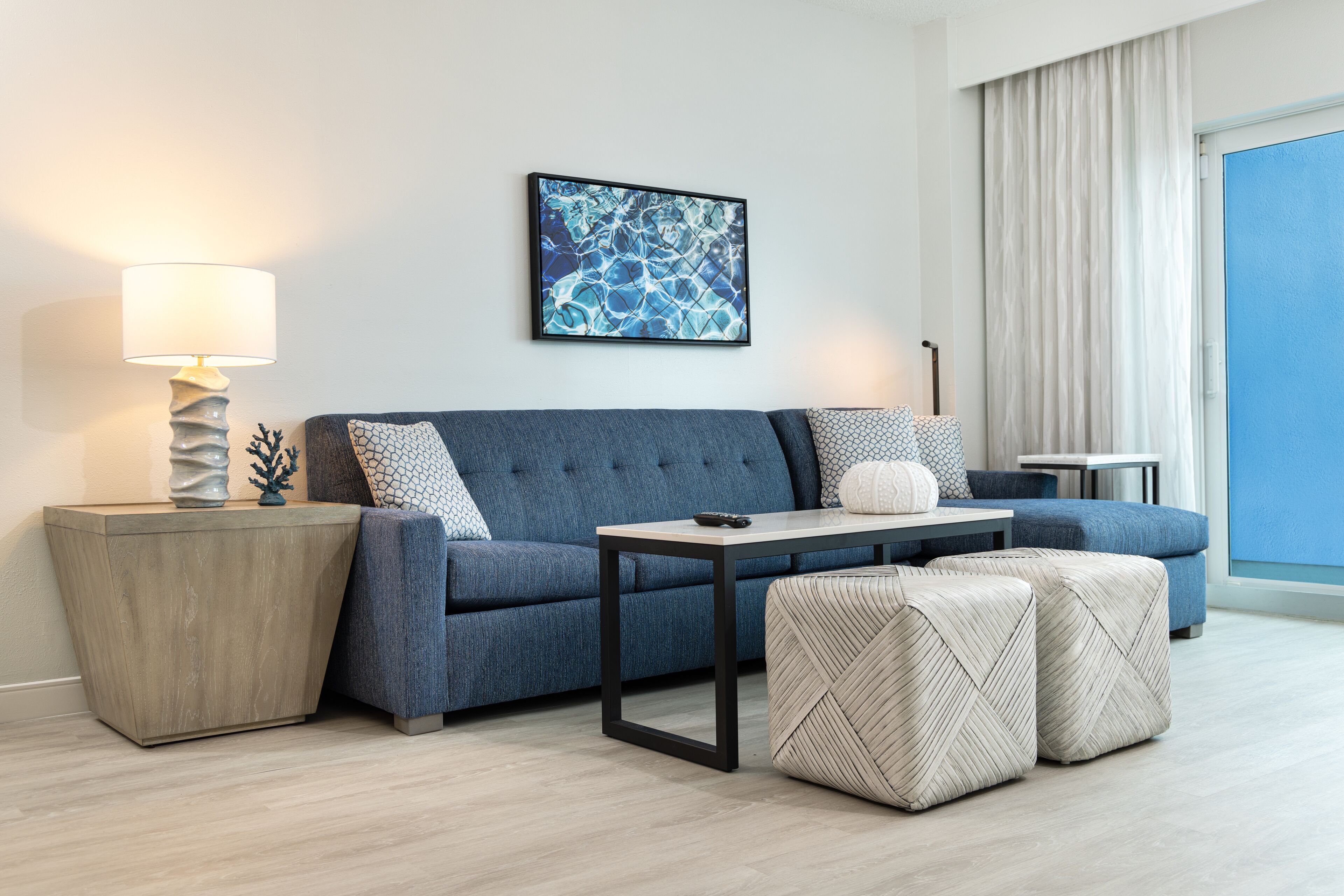 Una sala moderna con un sofá azul, una mesita de noche con una lámpara, una obra de arte abstracta enmarcada y vistas al exterior a través de un amplio ventanal.