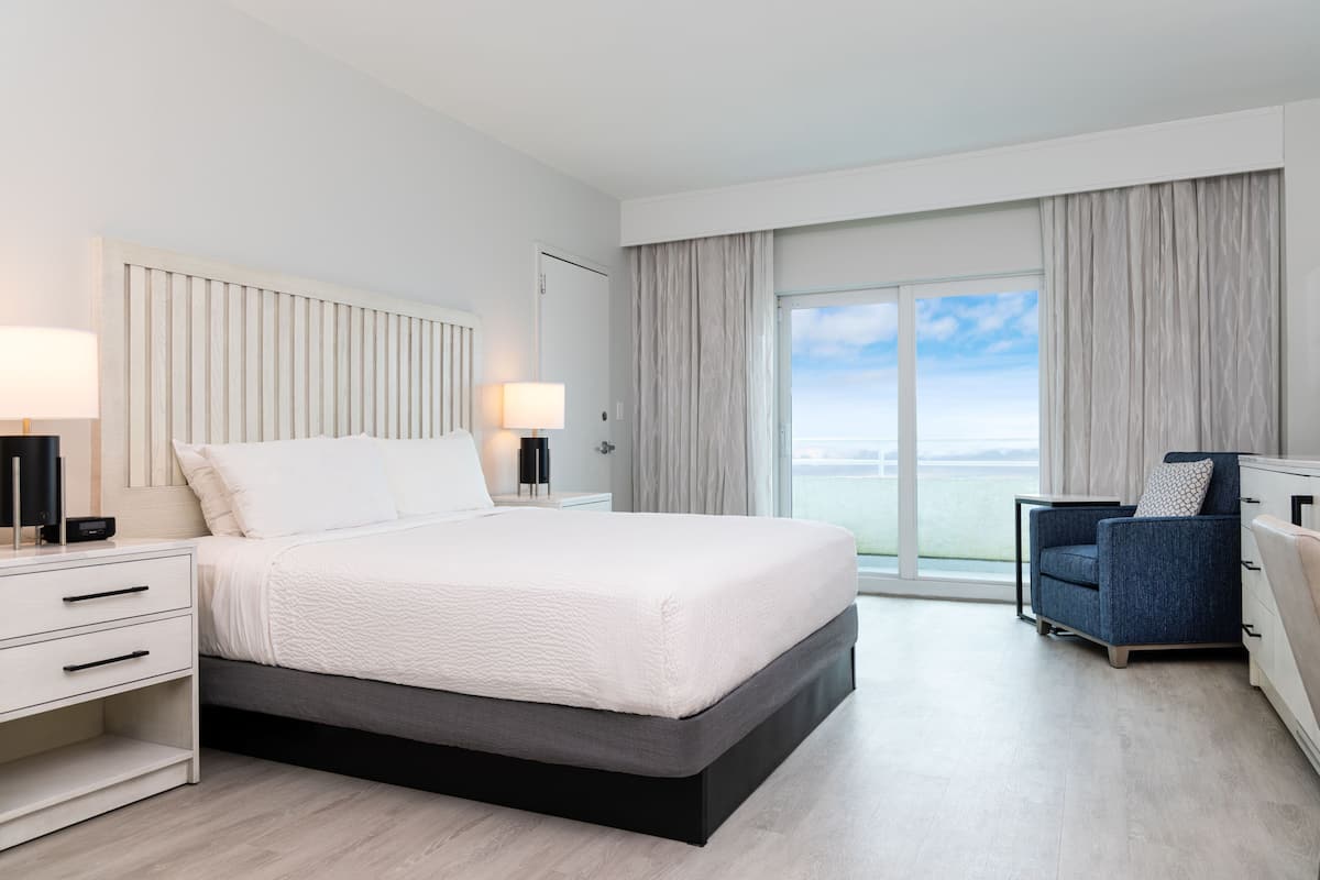 A modern hotel room with a large bed, a nightstand, a chair, and a view of the sea through the window.