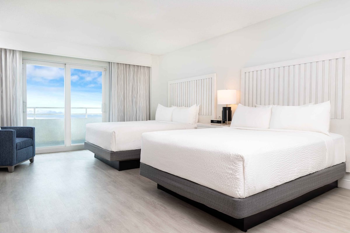 A hotel room with two beds, a chair, a large window with curtains, and a view of the sky.