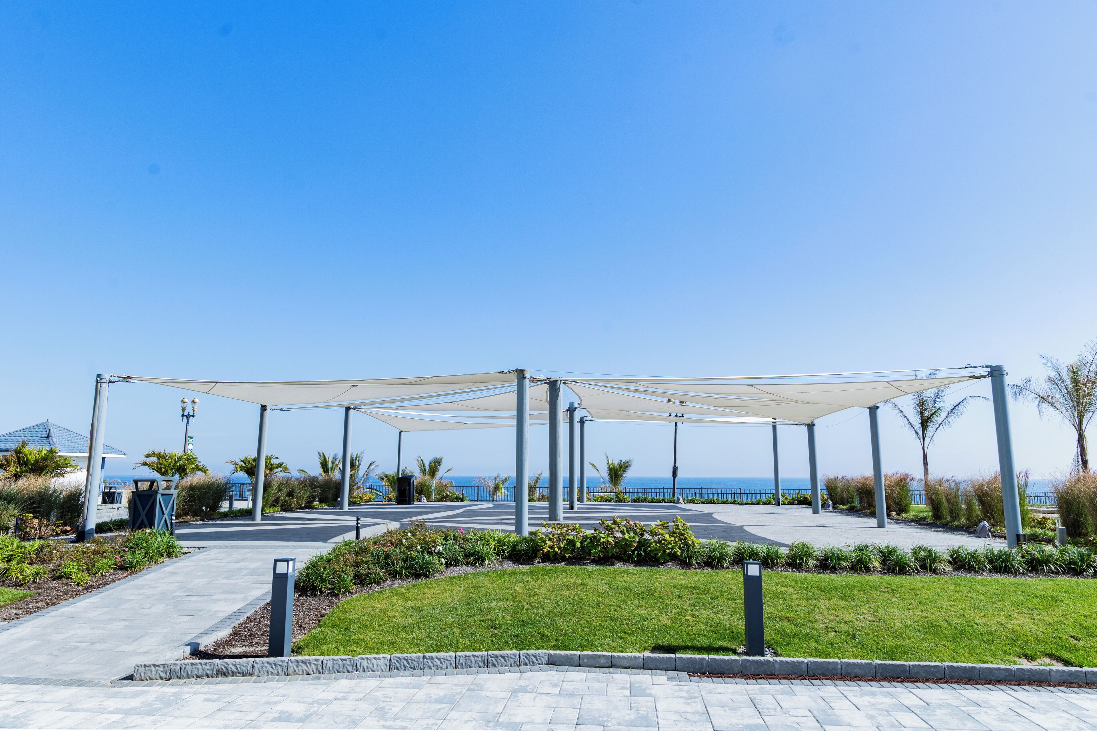A covered outdoor area with a pathway leading to a building, surrounded by greenery and a view of the sea.