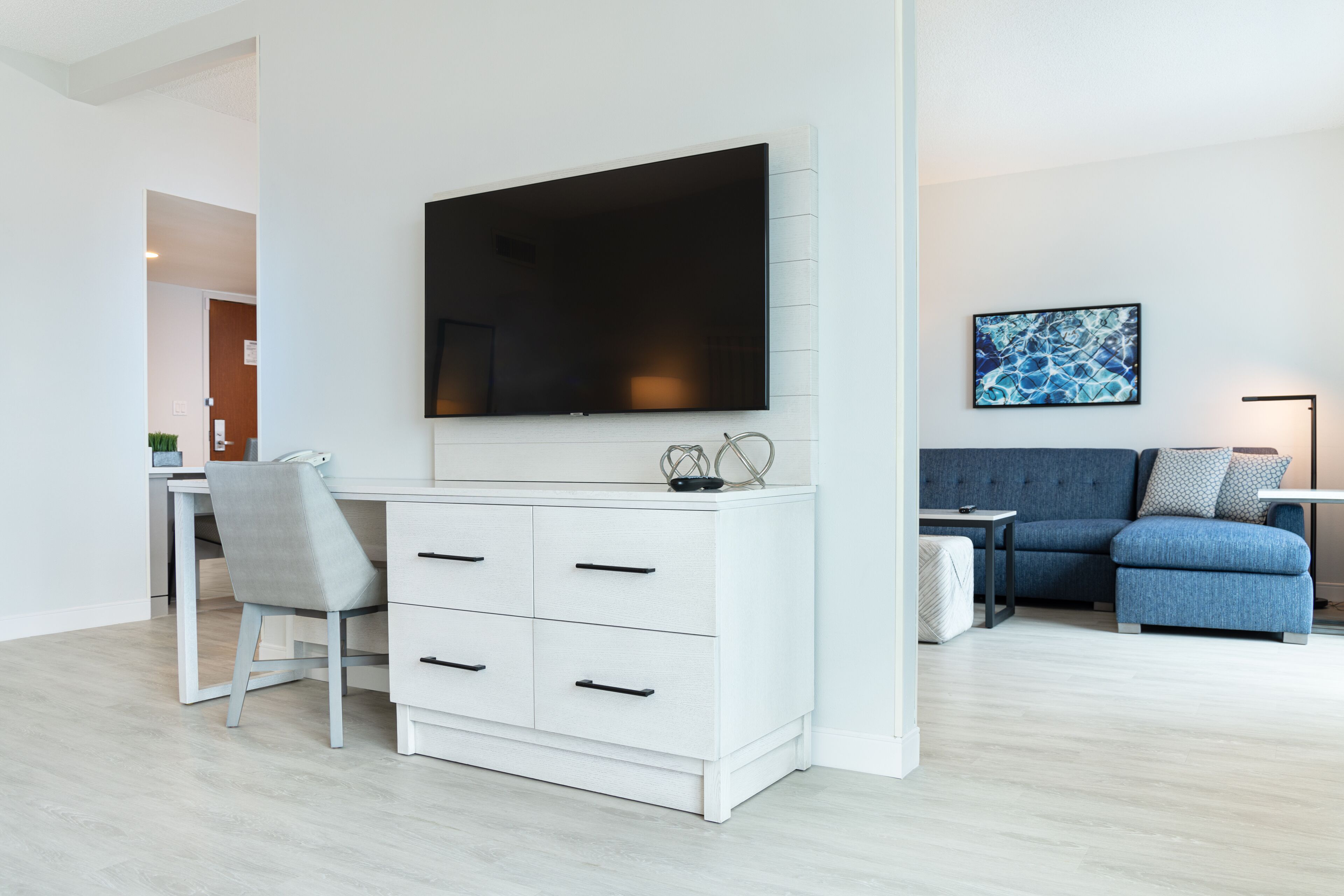 A modern living room with a white desk, a flat-screen TV mounted on the wall, a blue sofa, and a framed abstract painting.