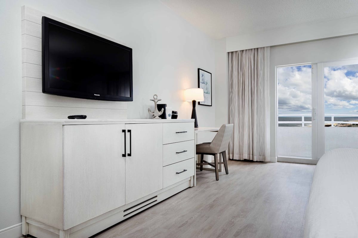 A hotel room with a large flat-screen TV mounted on the wall, a desk with a chair, a bed, and a large window with a view of the sea.