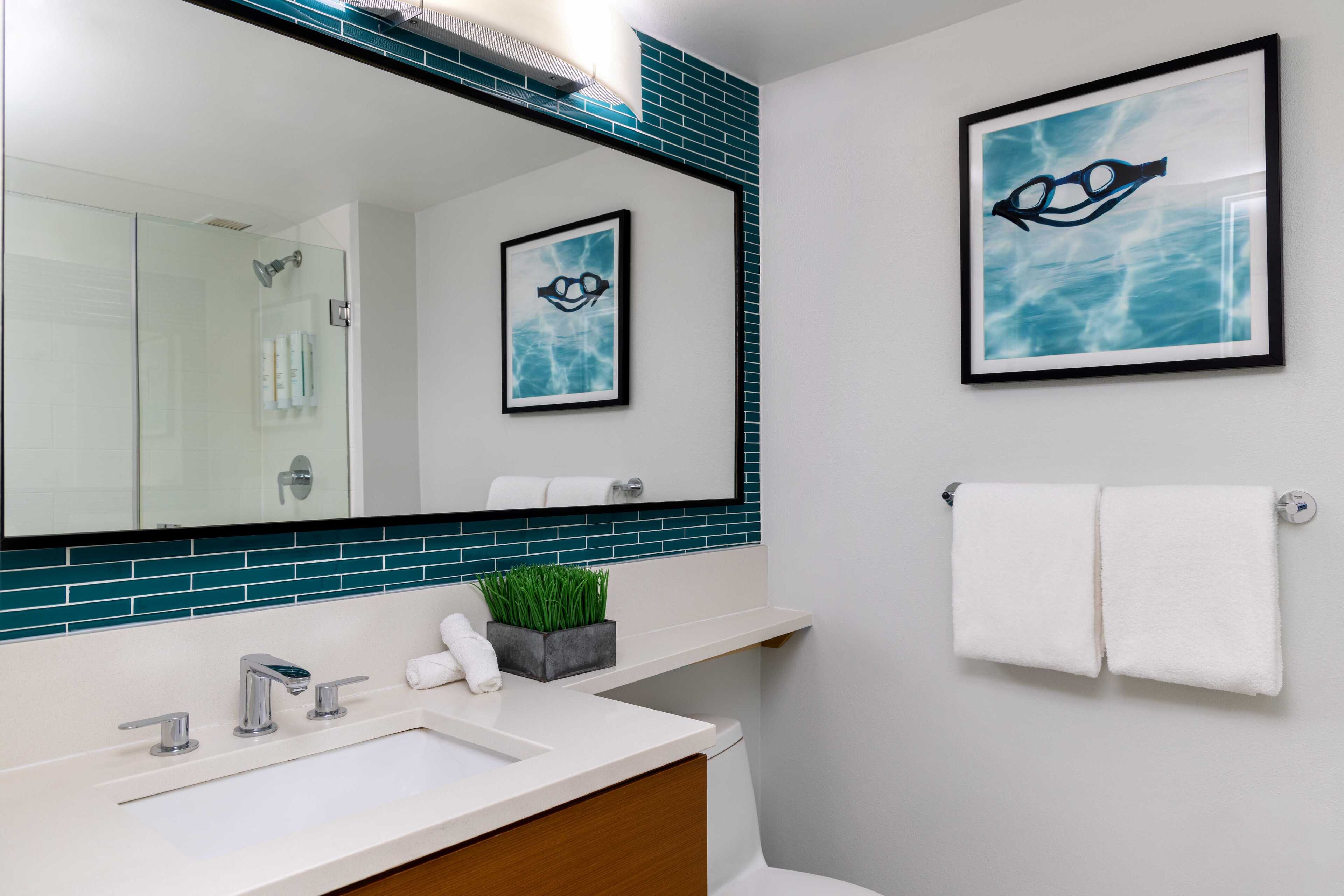 A bathroom with a large mirror, a white sink, a towel rack, and a framed picture of swimming goggles.