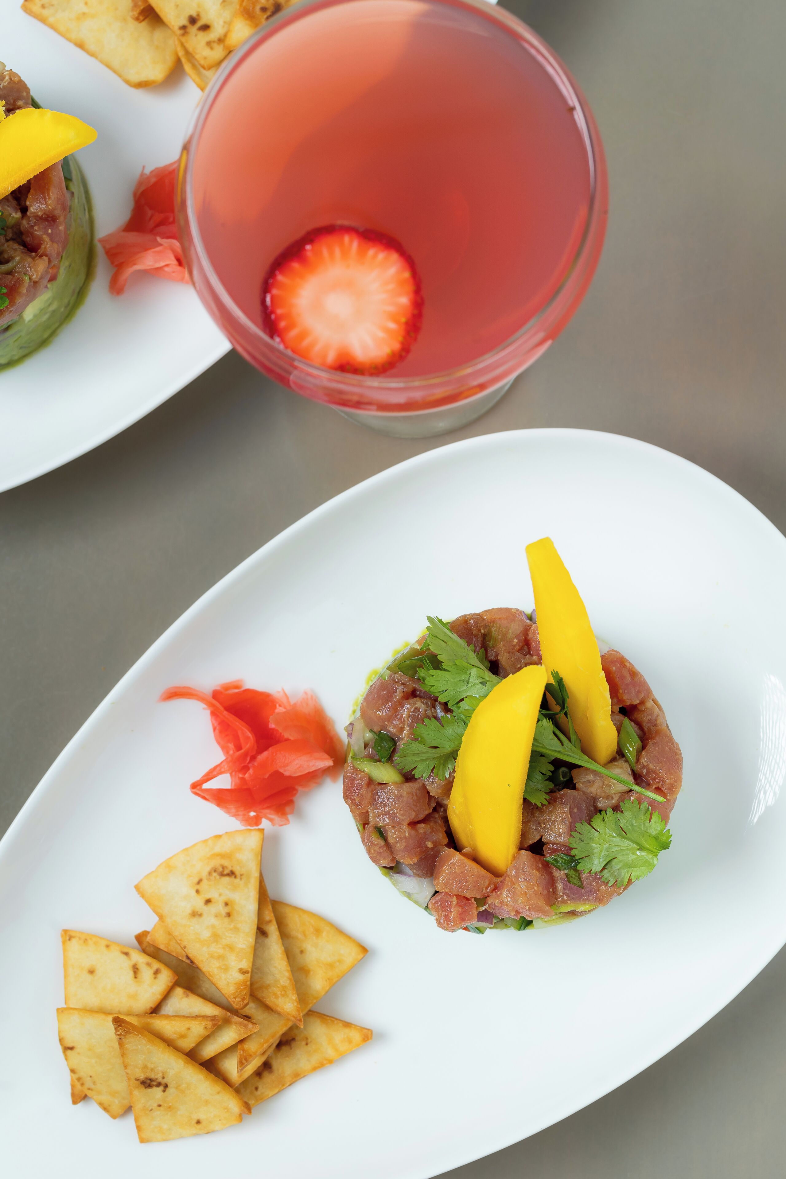 A plate with a dish of raw fish, garnished with yellow and red elements, and a glass of pink beverage with a strawberry slice.