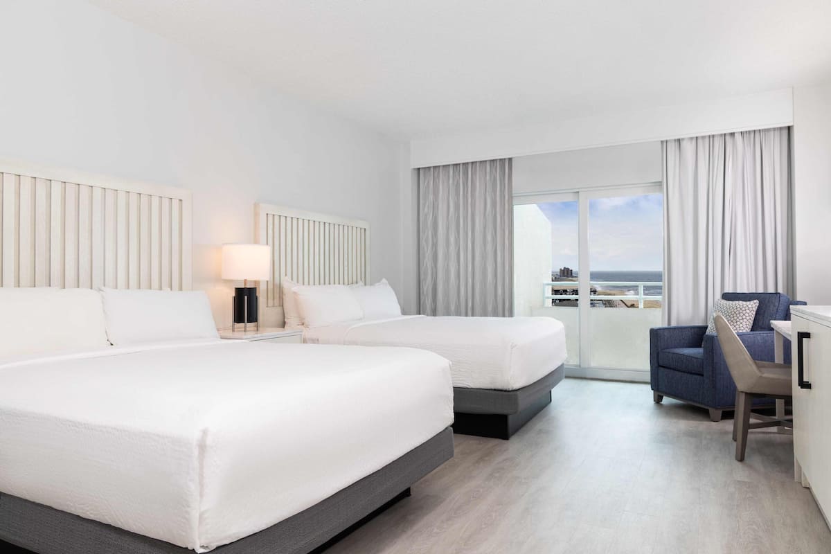 A hotel room with two beds, a desk, and a view of the sea.