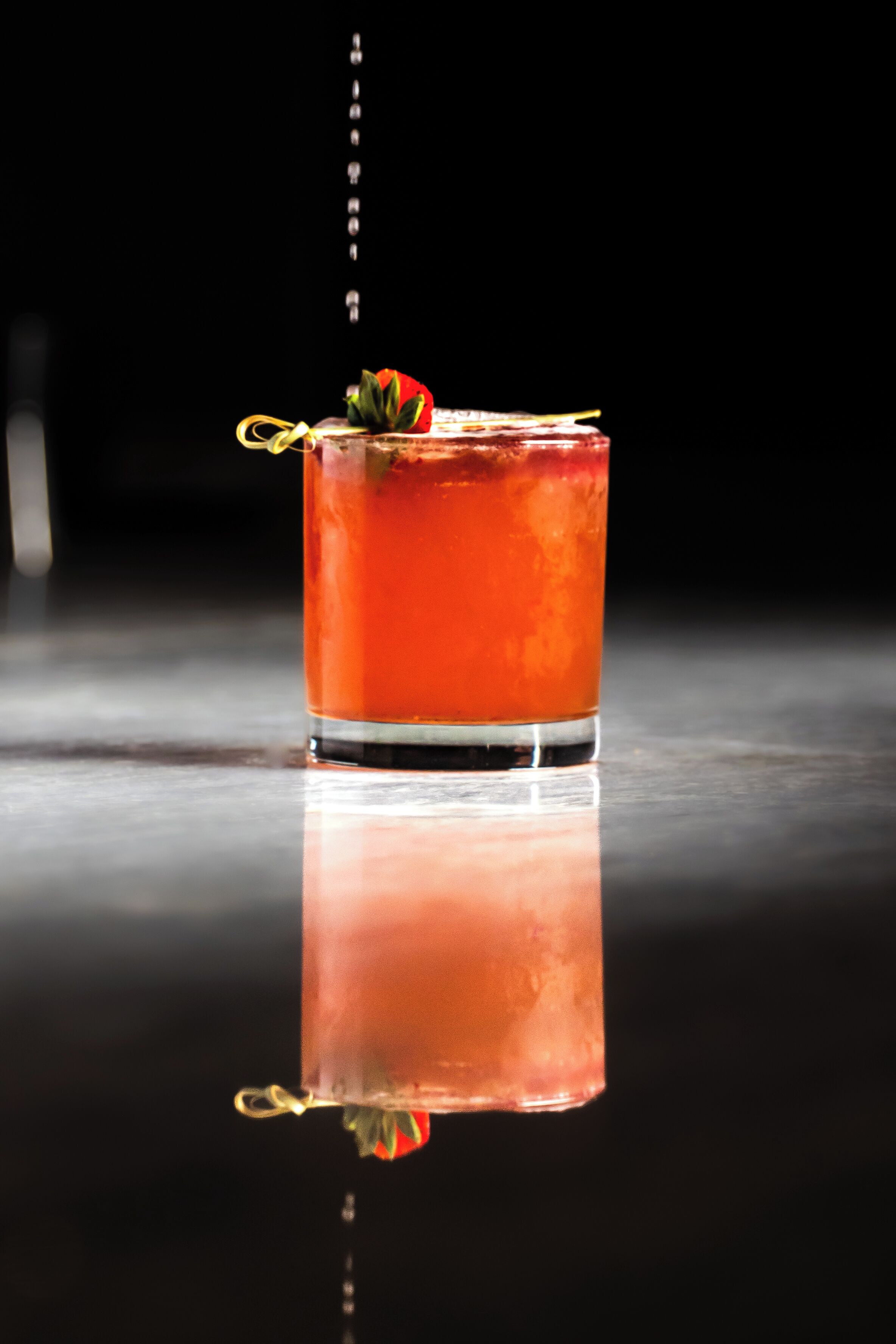 A clear glass of red beverage with a strawberry garnish on a reflective surface.