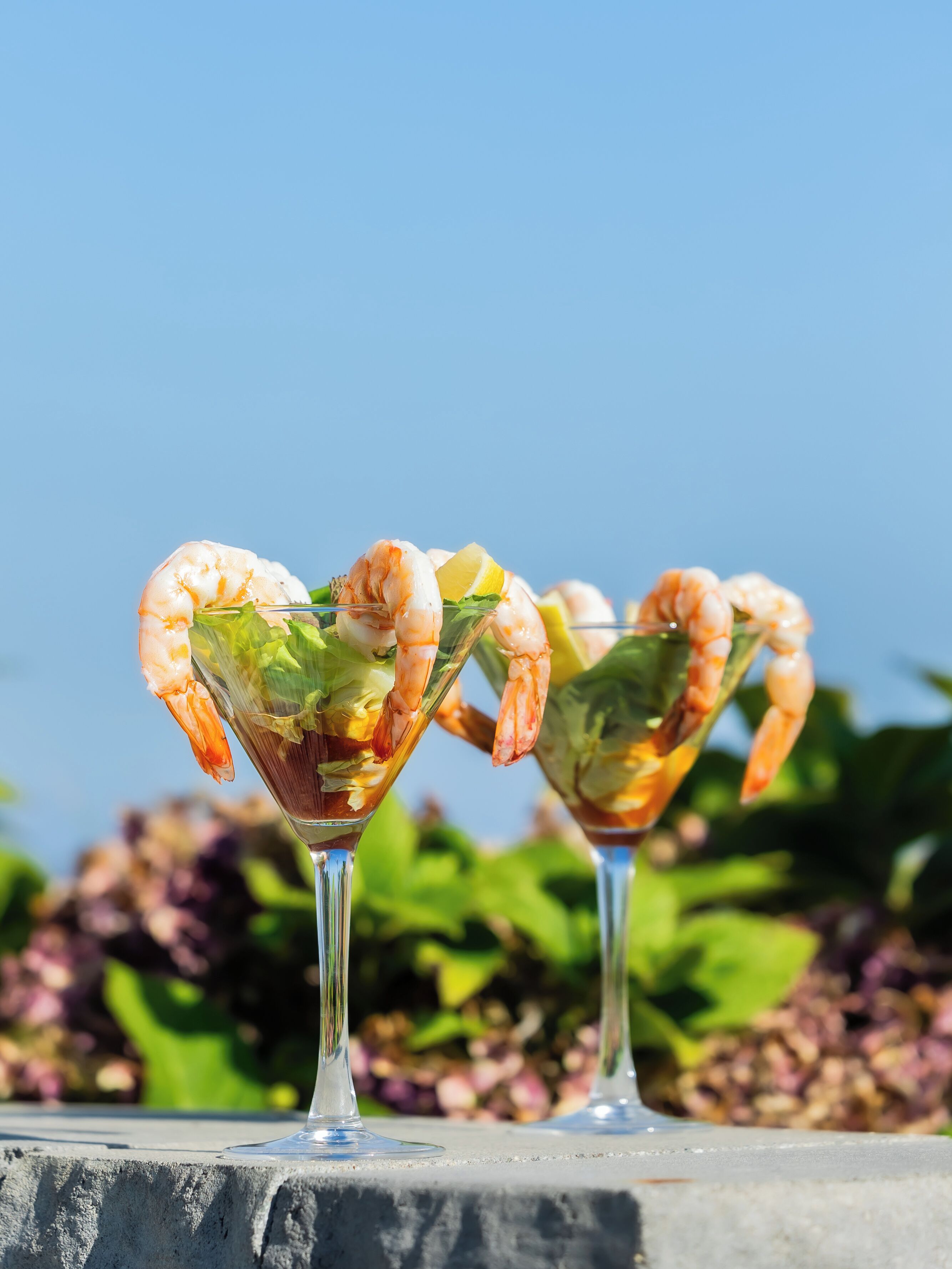 Two martini glasses filled with shrimp cocktails on a concrete surface with a blurred garden background.