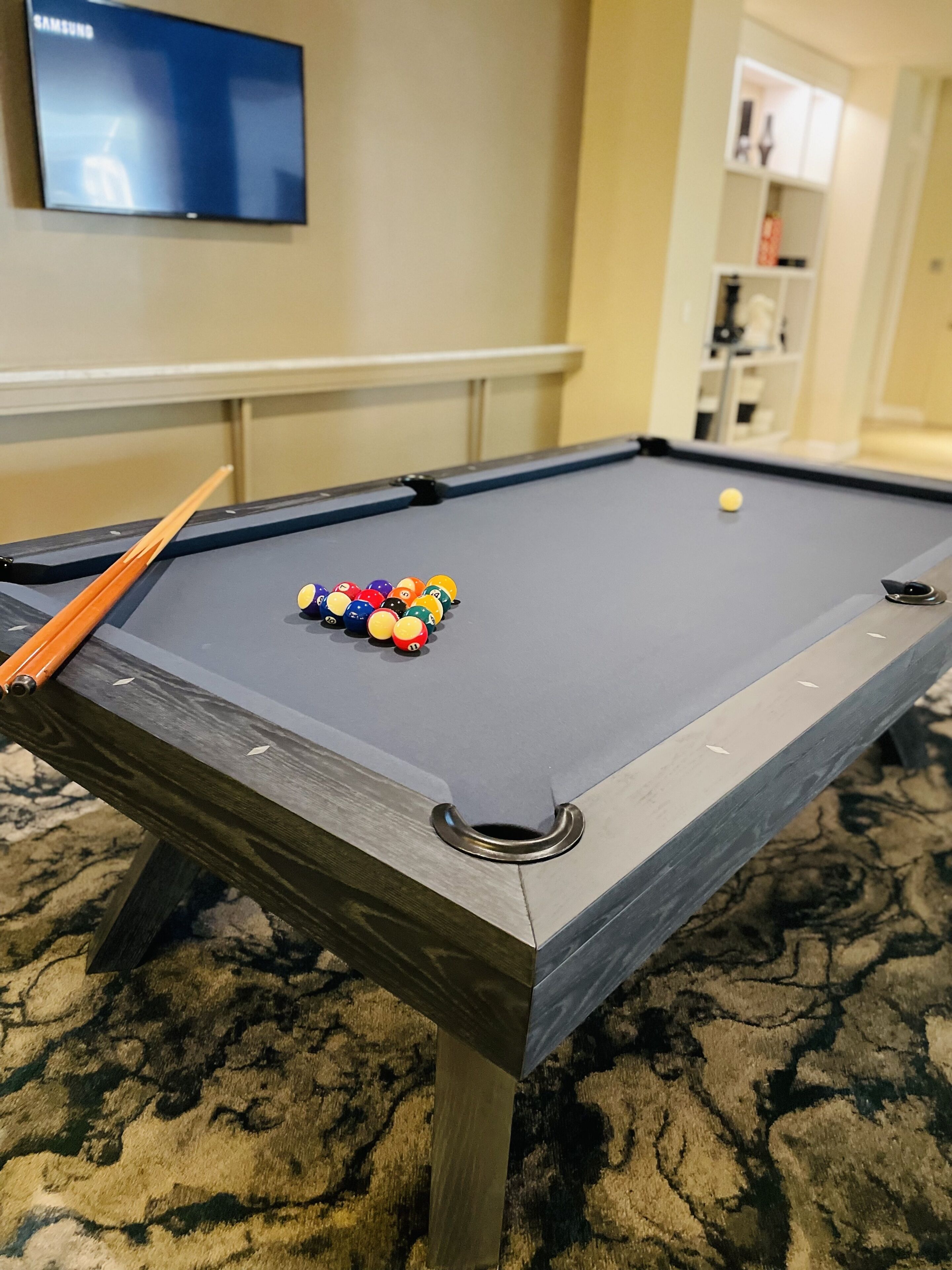 A pool table with a cue stick and billiard balls, a television on the wall, and a carpeted floor.