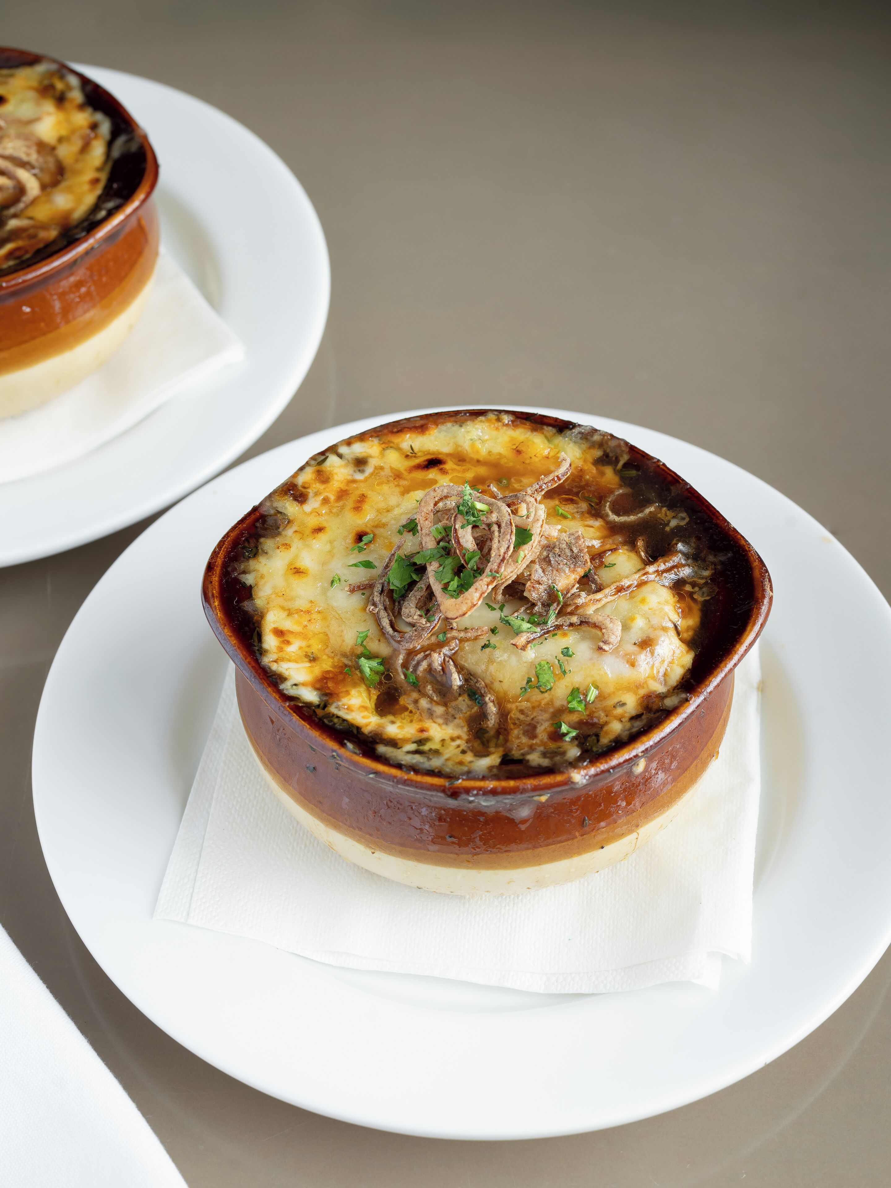 Un plato de papas al horno cubiertas de queso y cebollas, servido en un recipiente de cerámica marrón sobre un plato blanco.