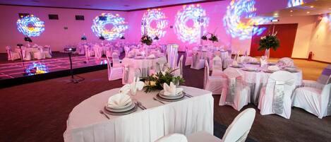 Un salón de banquetes con mesas redondas dispuestas para un evento formal, con manteles blancos, cubertería y centros de mesa florales.