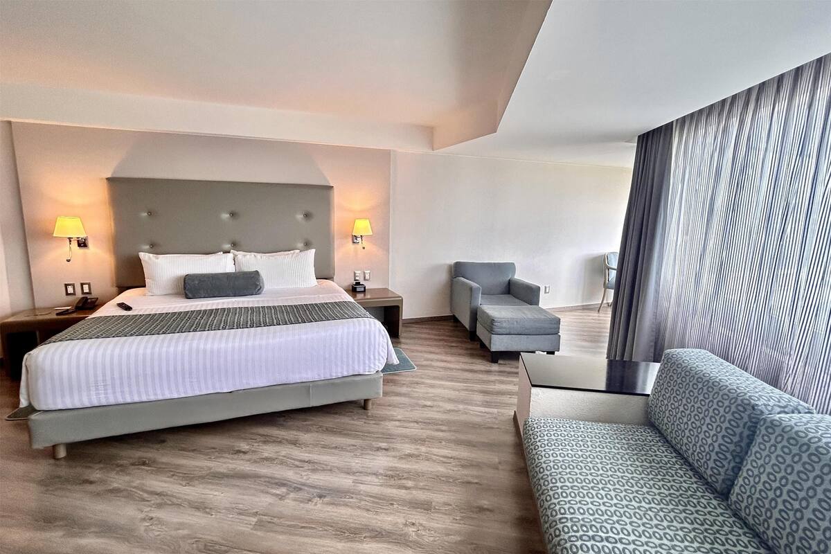 A hotel room with a large bed, two bedside lamps, a grey armchair, a patterned sofa, and a large window with curtains.