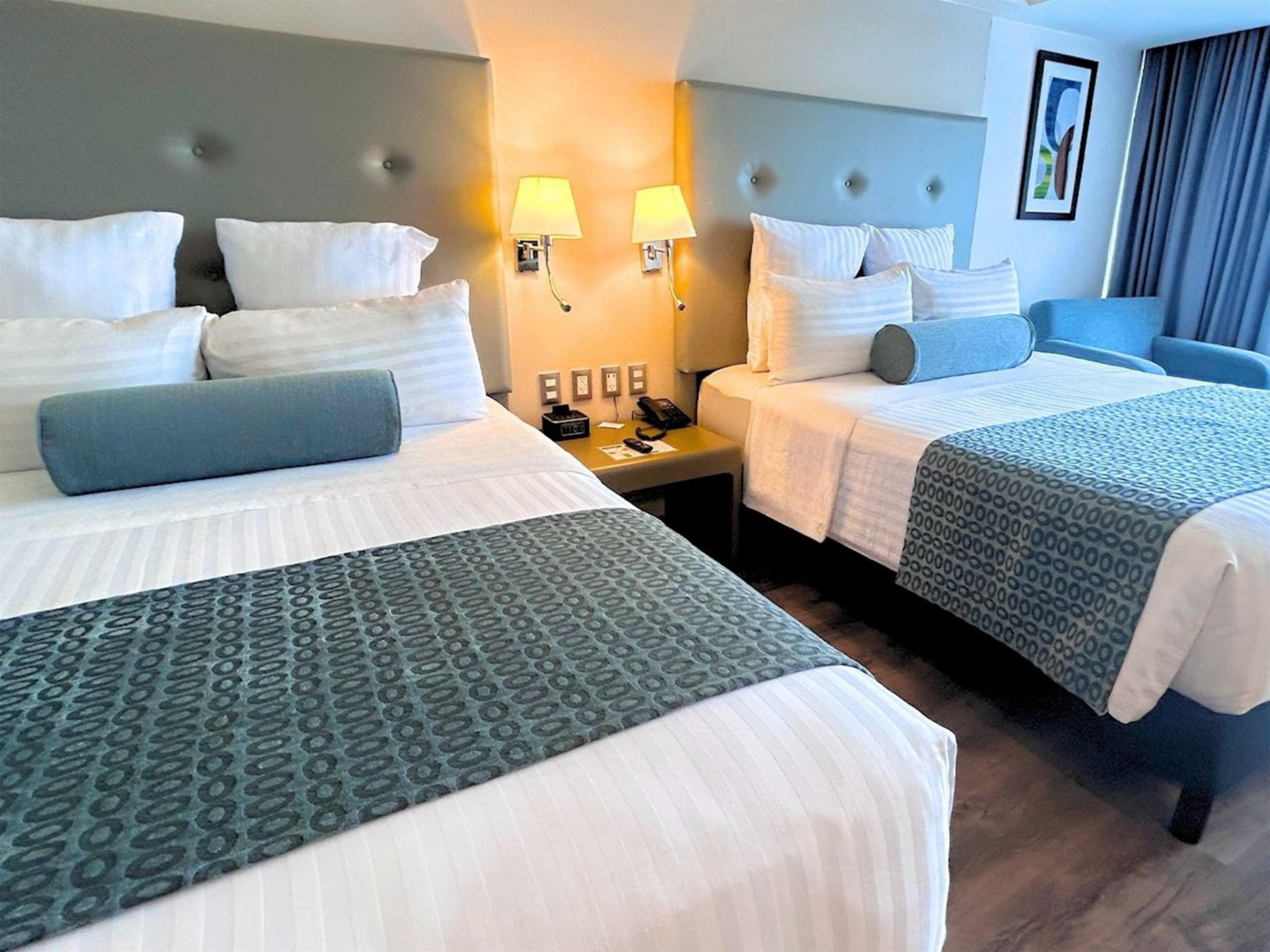 A hotel room with two beds, each with white linens and blue accents, a bedside table with a lamp, and a wall-mounted phone.