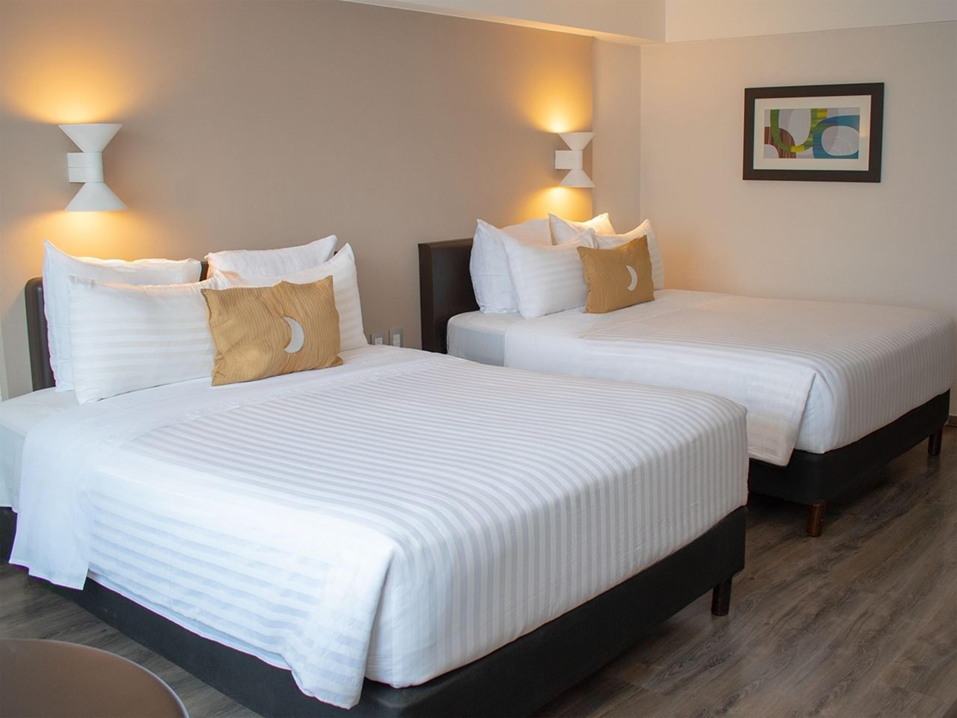 A hotel room with two beds, each with white bedding and a moon-shaped pillow. There are wall-mounted light fixtures on either side of the beds.