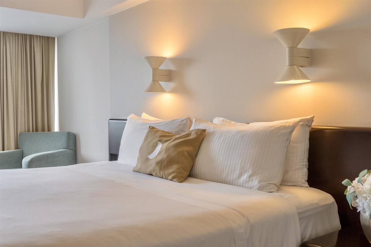 A hotel room with a large bed, two wall-mounted light fixtures, a grey armchair, and a vase with flowers.