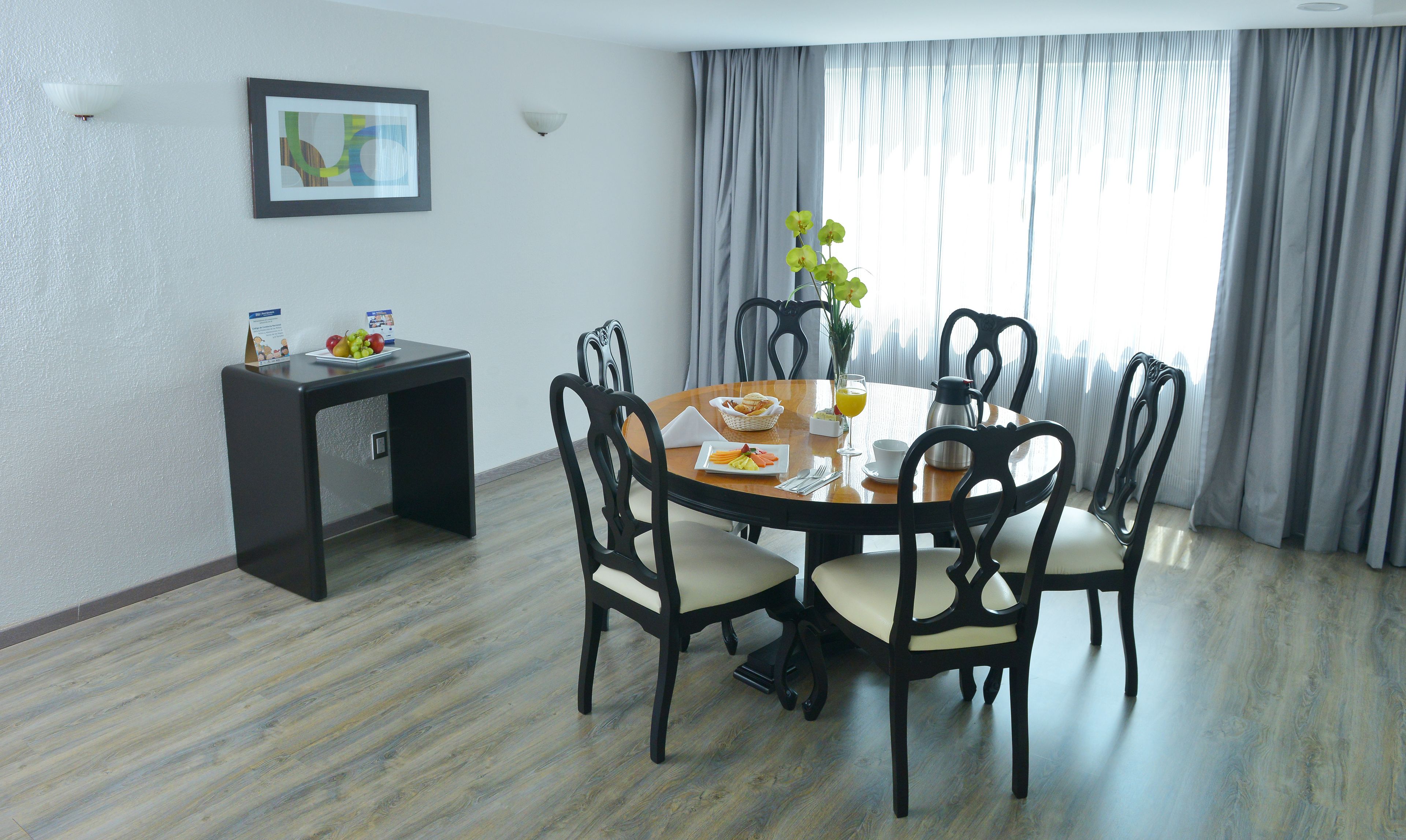 A dining area with a round table, six chairs, a small side table with fruits, a framed picture on the wall, and a window with curtains.