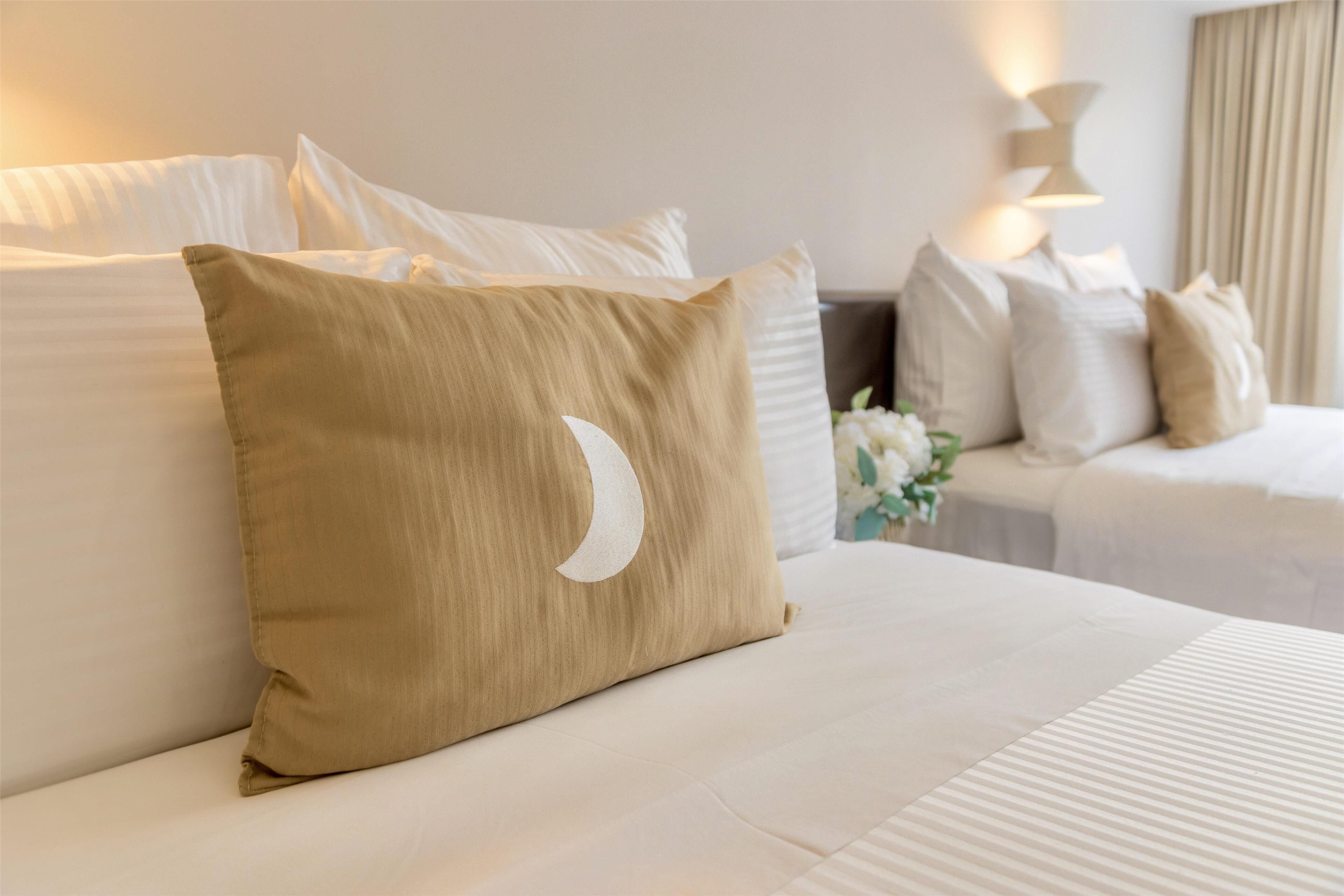 A hotel room with two beds, each with white linens and multiple pillows, including a beige pillow with a crescent moon design.